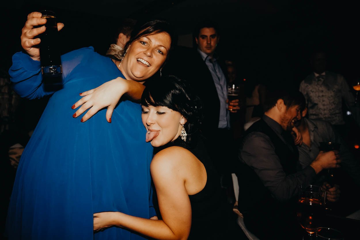 Two women enjoying a fun moment at a lively indoor social wedding, one woman in blue dress hugging and holding a drink, the other in black dress with her tongue out, sitting with others in a dimly lit, celebratory wedding in Bridgend, south wales. Sh