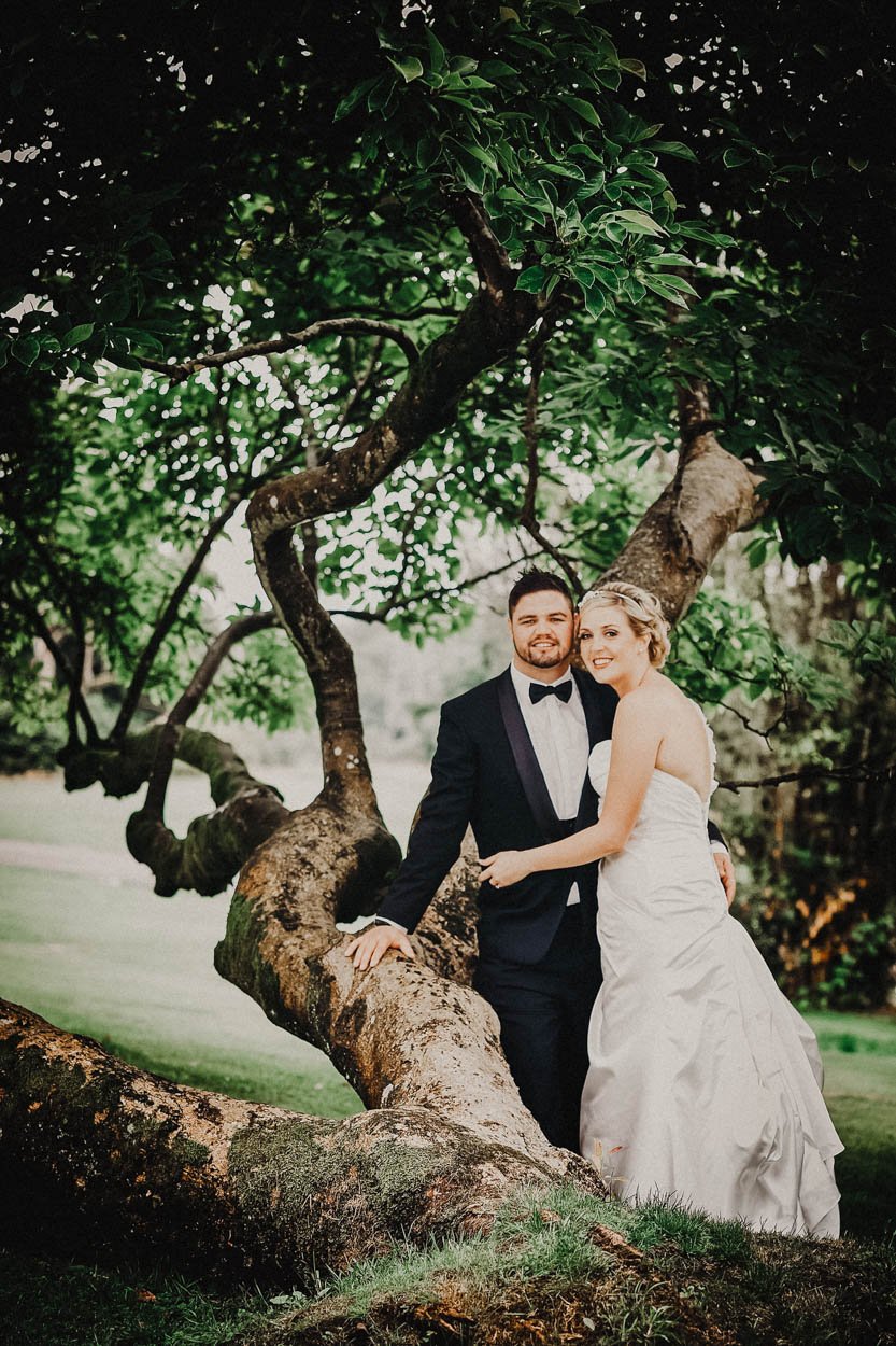 A bride and groom posing outdoors next to a large, twisted tree with green leaves on a cloudy day in Court colman hotel, Bridgend. Shot by Jason Evans photography Bridgend