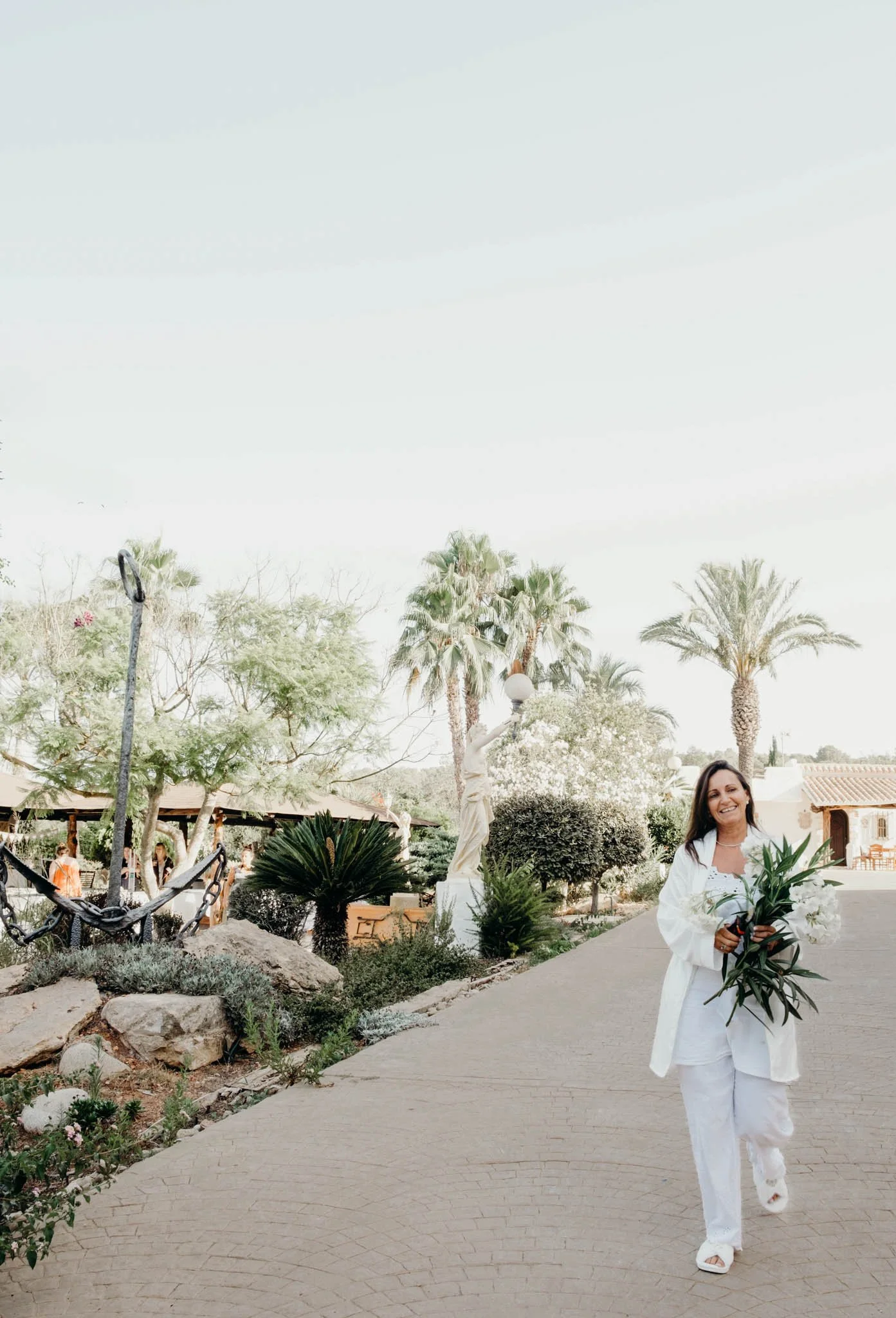 A woman in white clothing smiling and holding a bouquet of white flowers, walking along a paved path in a tropical garden with palm trees.