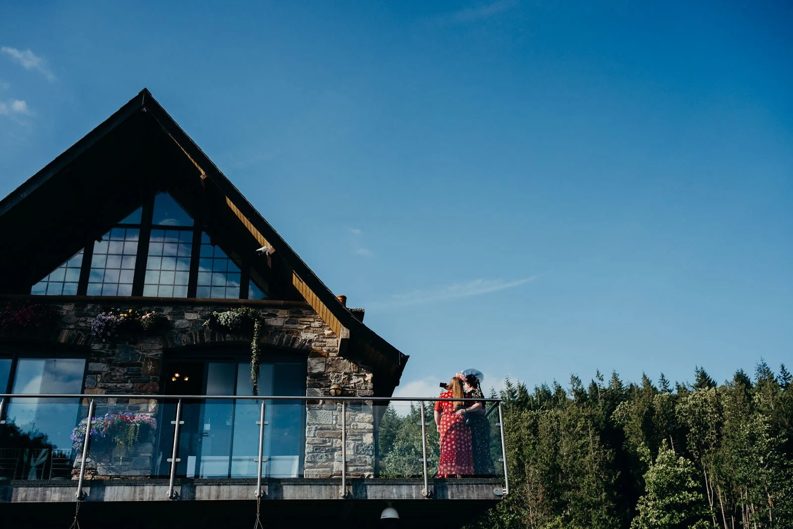 Two women standing on a balcony in front of a house with a stone facade and large windows, with trees and a blue sky in the background.