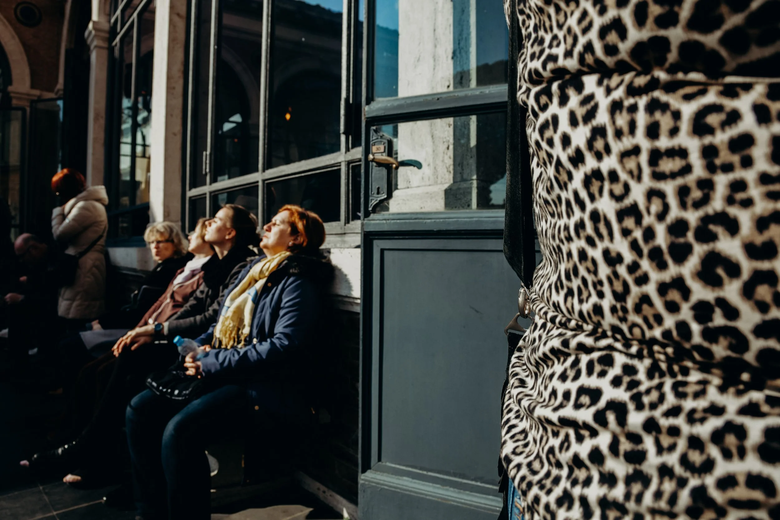 Three women sit on a bench outdoors in sunlight, eyes closed and heads tilted upward enjoying the warmth. In the foreground, a leopard-printed woman partially obscuring the view