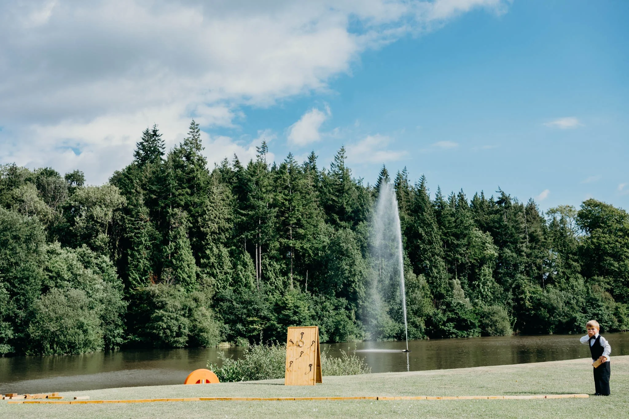 A young boy dressed in formal attire standing on a grassy area near a lake, with a large fountain spraying water into the air in the background, surrounded by trees and a partly cloudy sky at Canada lake lodge pentyrch. 