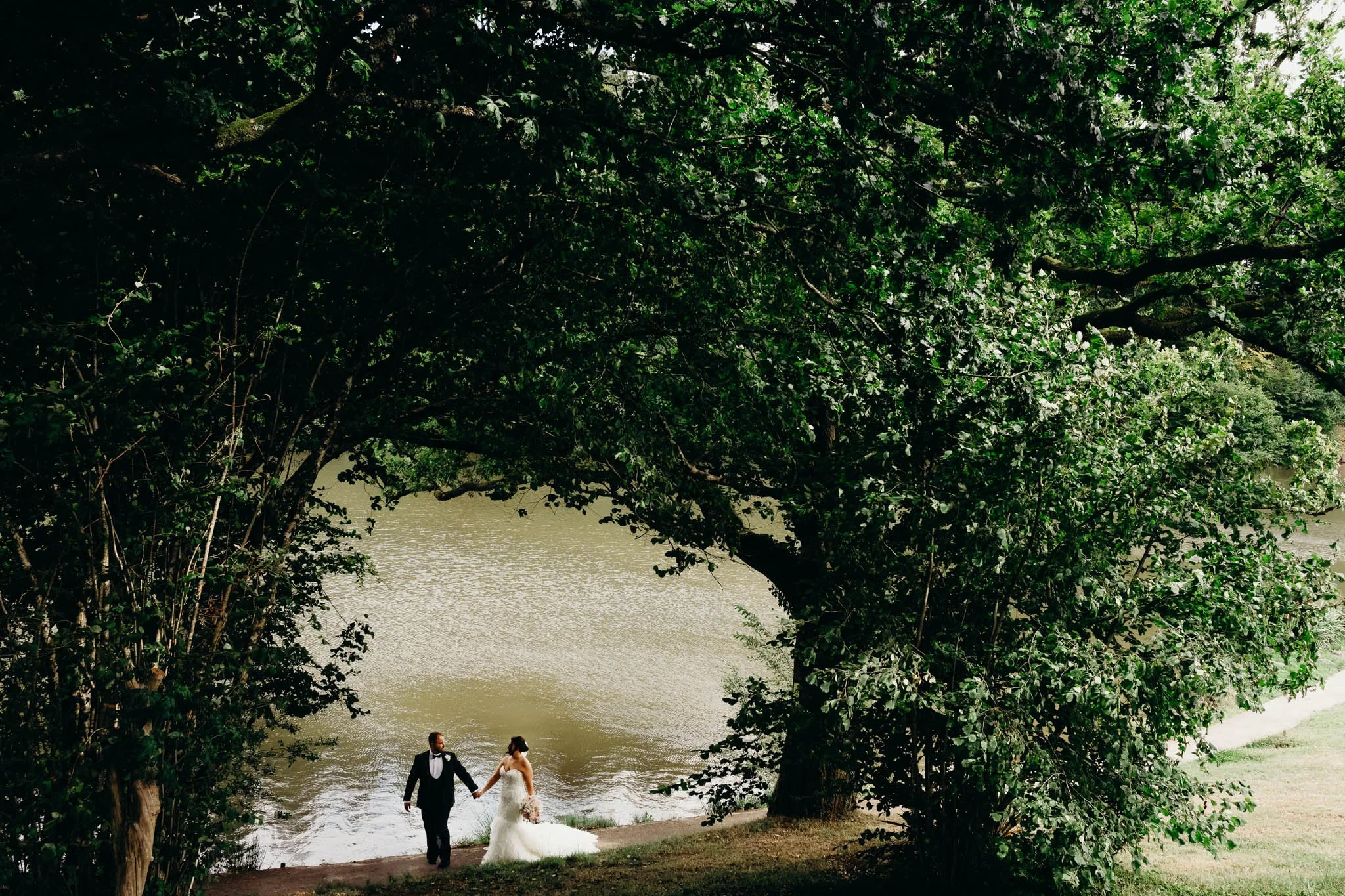 A bride and groom holding hands by a body of water under a large tree with dense green foliage at Canada lake lodge pentyrch. 