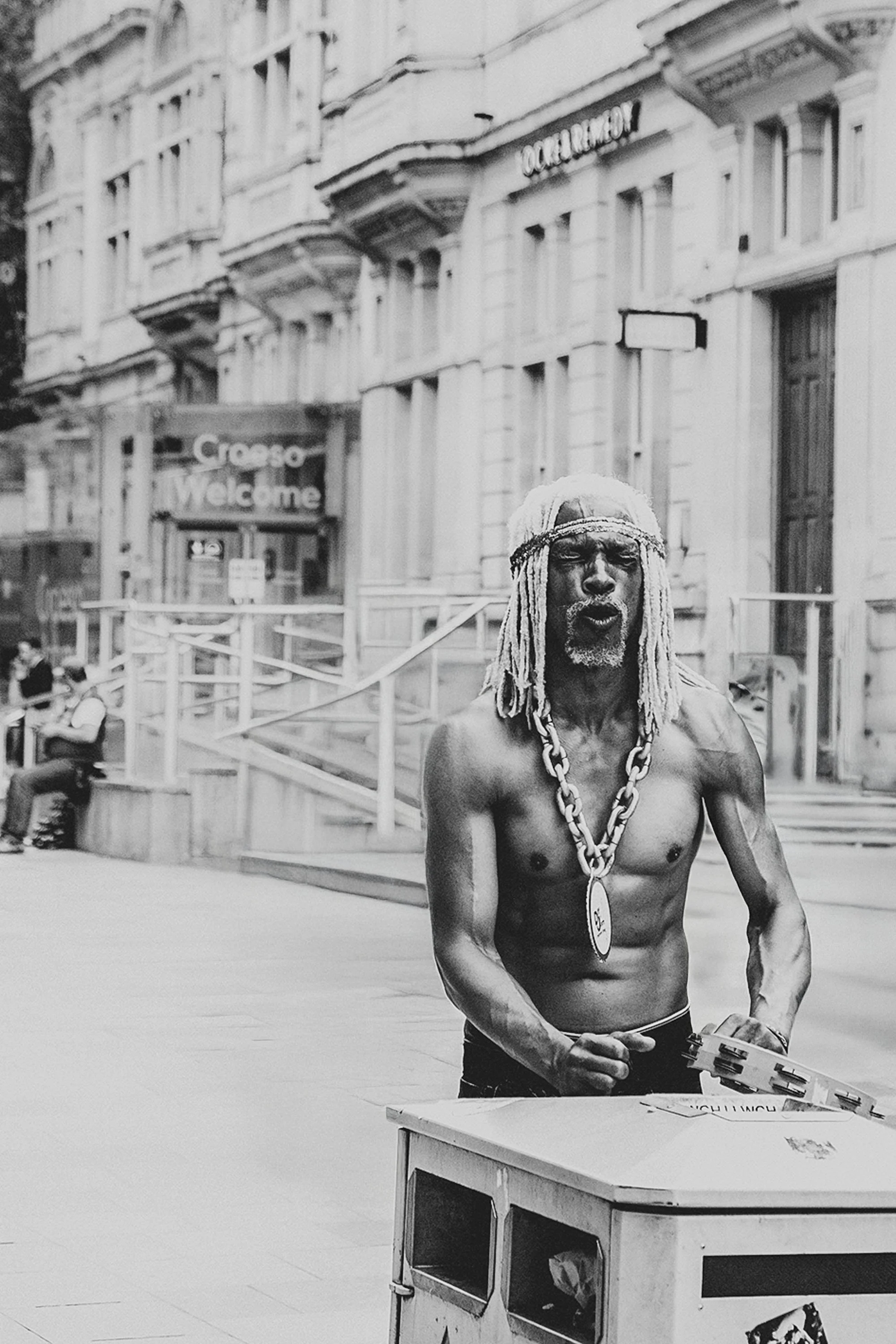 A muscular, shirtless man with long braided hair, a headband, and a large chain necklace stands behind a bin on the streets of Cardiff singing. 