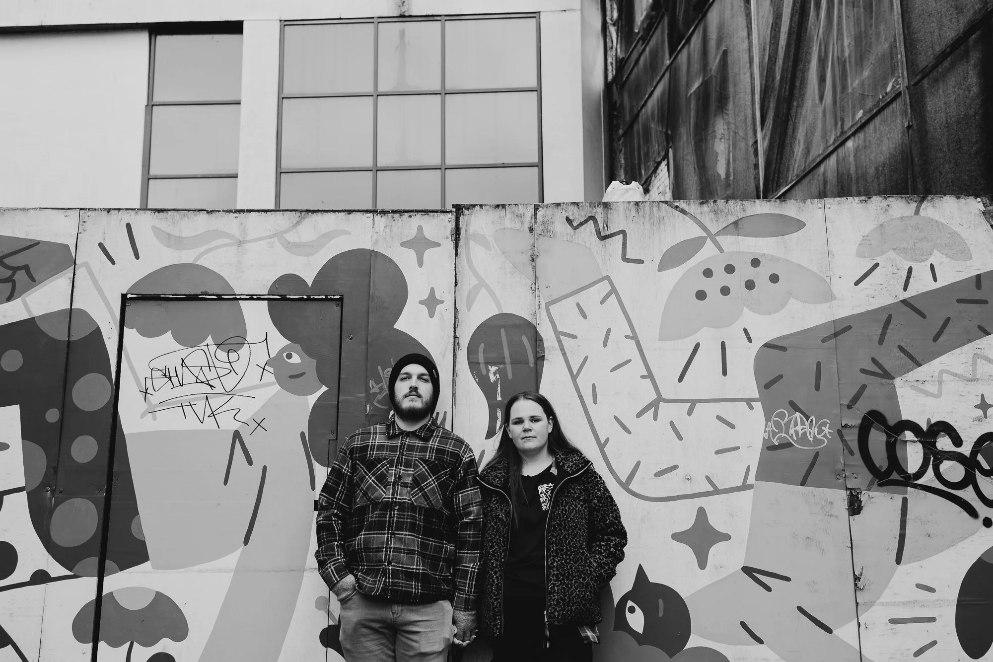 Black and white photo of a couple holding hands back-to-back against a graffiti mural, fun city engagement photo