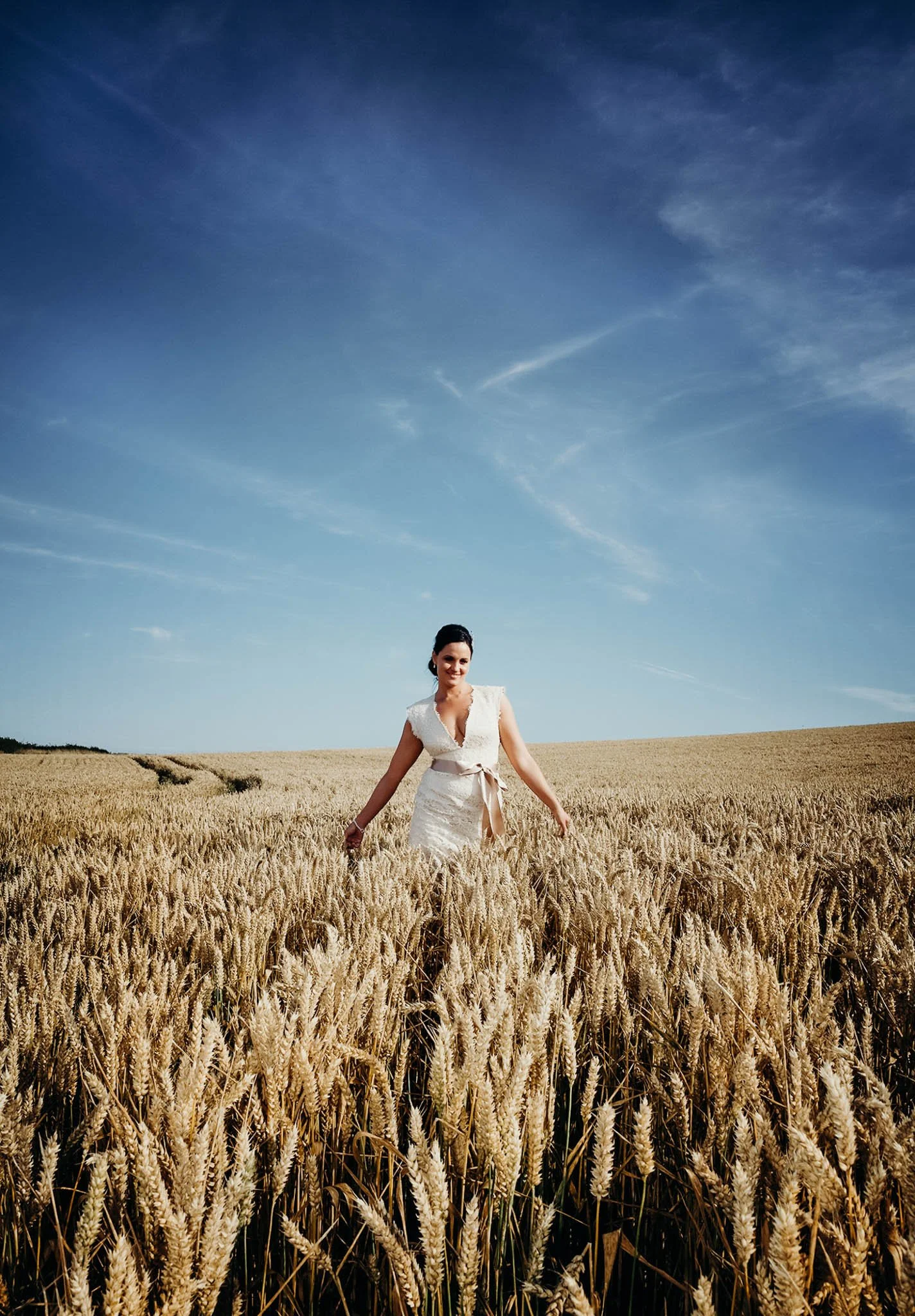 A woman in a white dress walking through a golden wheat field under a blue sky with wispy clouds.