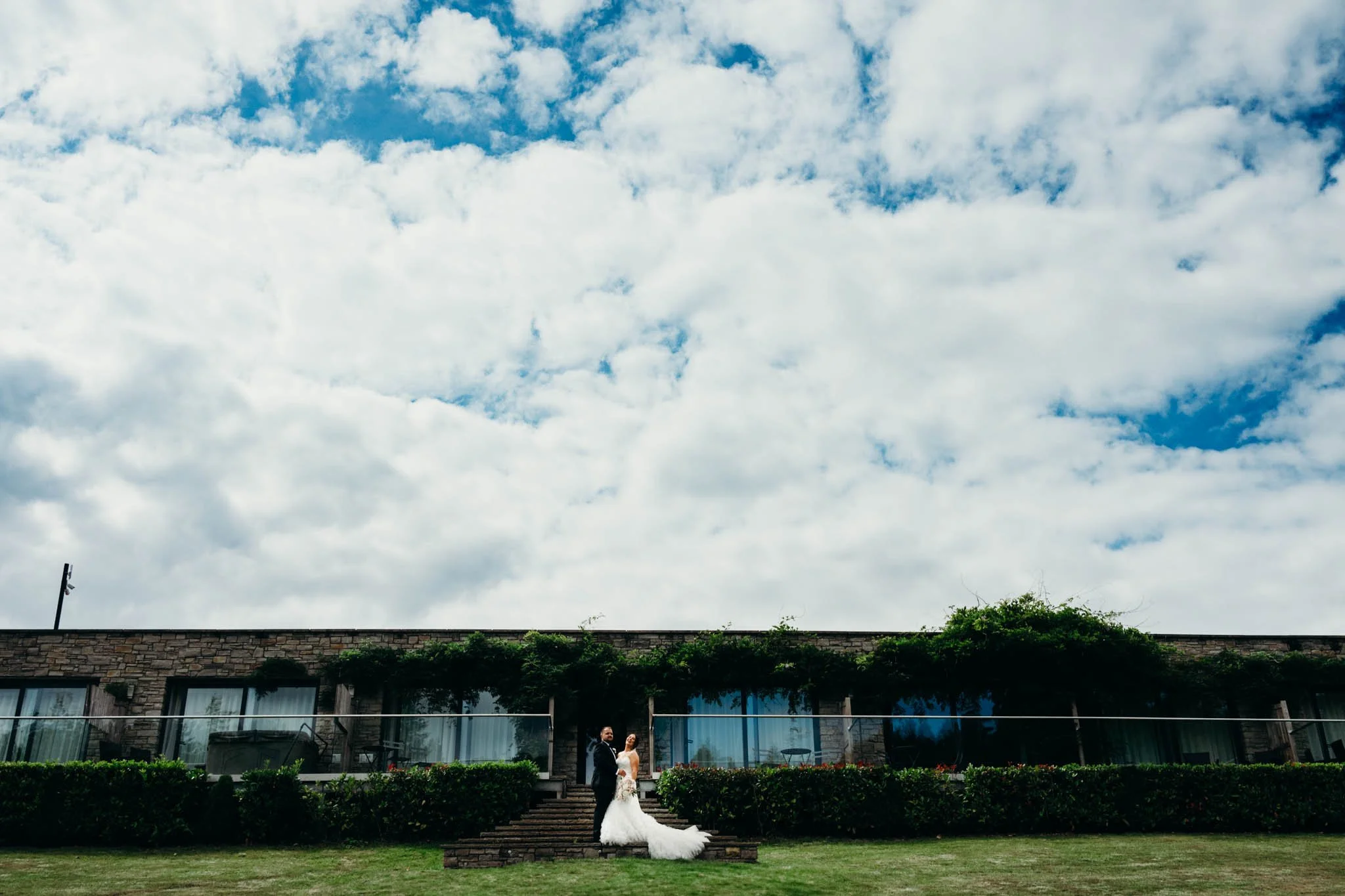 A bride and groom in wedding attire standing on brick stairs outside a modern building with large glass windows and greenery on the roof, with a partly cloudy sky overhead at Canada lake lodge pentyrch. 