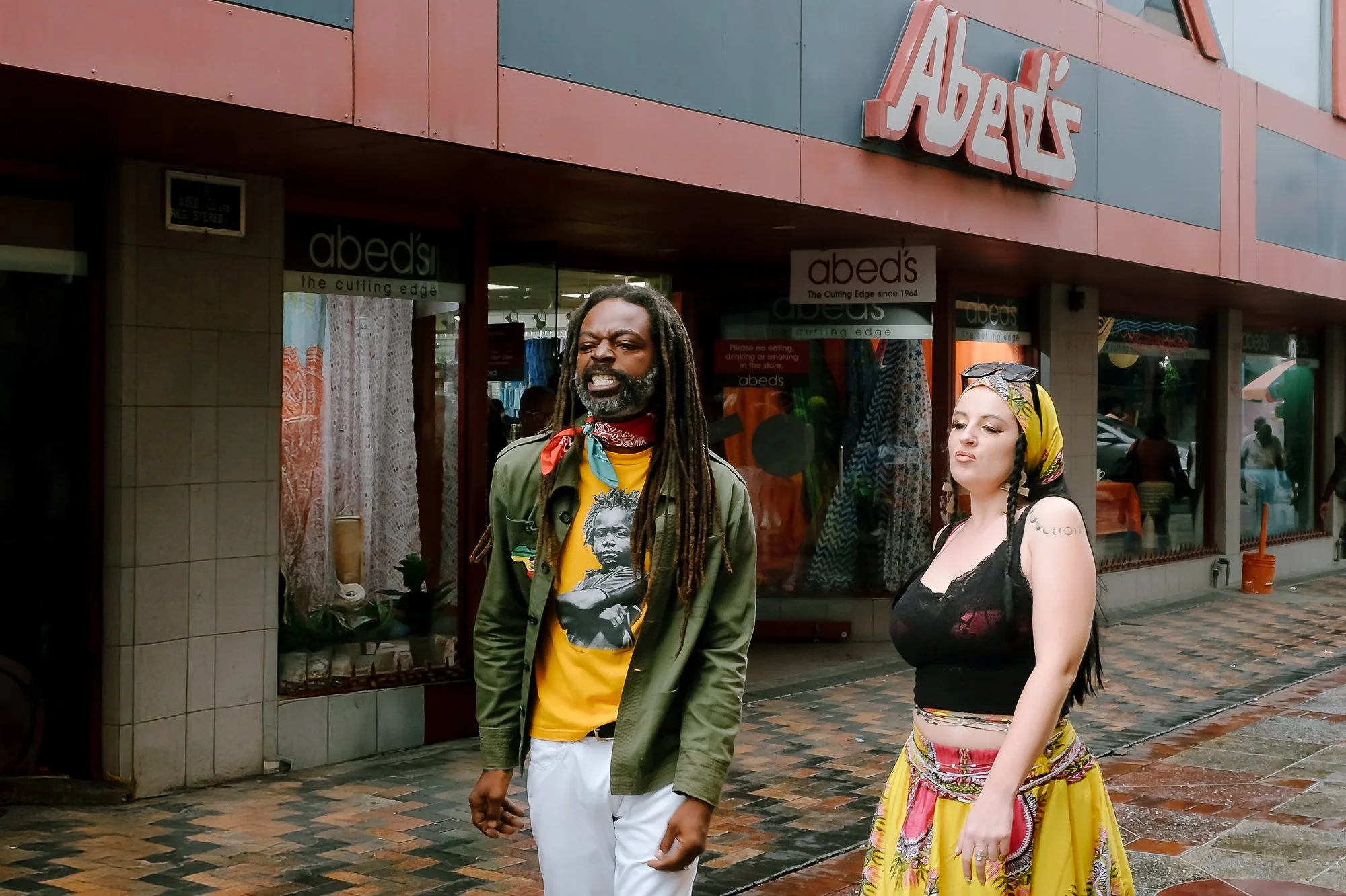In Bridgetown, Barbados, a man with long dreadlocks wearing a yellow T-shirt with a portrait print, red bandana, and green jacket walks alongside a woman in a colorful headscarf, black lace top, and vibrant skirt. They are in front of a storefront na