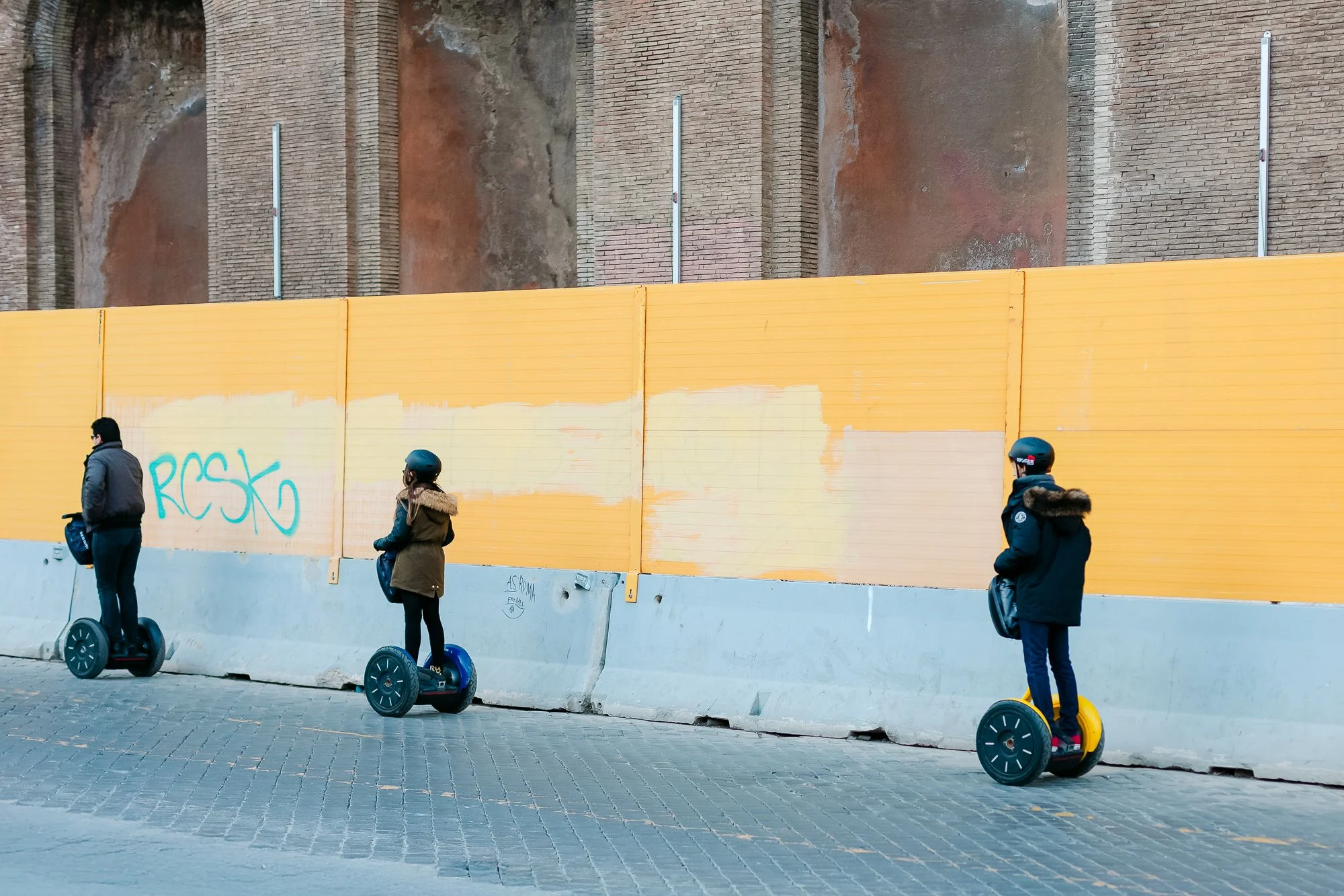 Three tourists wearing helmets ride two-wheeled self-balancing scooters (Segways) in a line on a cobblestone street in front of a tall orange construction barrier with graffiti reading "ROSKO." Behind the barrier is an ancient brick wall, likely part