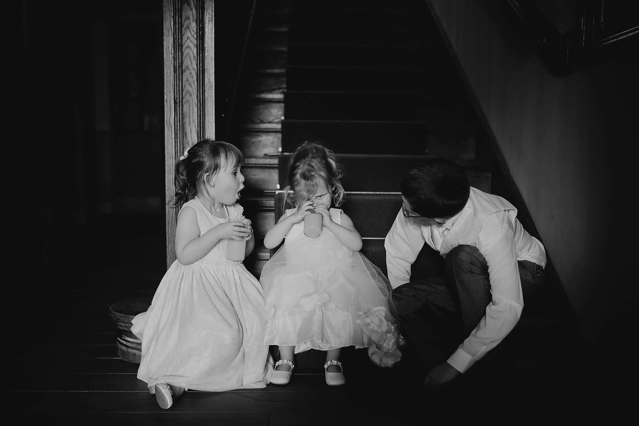 Three young girls and a young boy sitting on a wooden staircase, with two girls drinking from cups and a boy looking down.