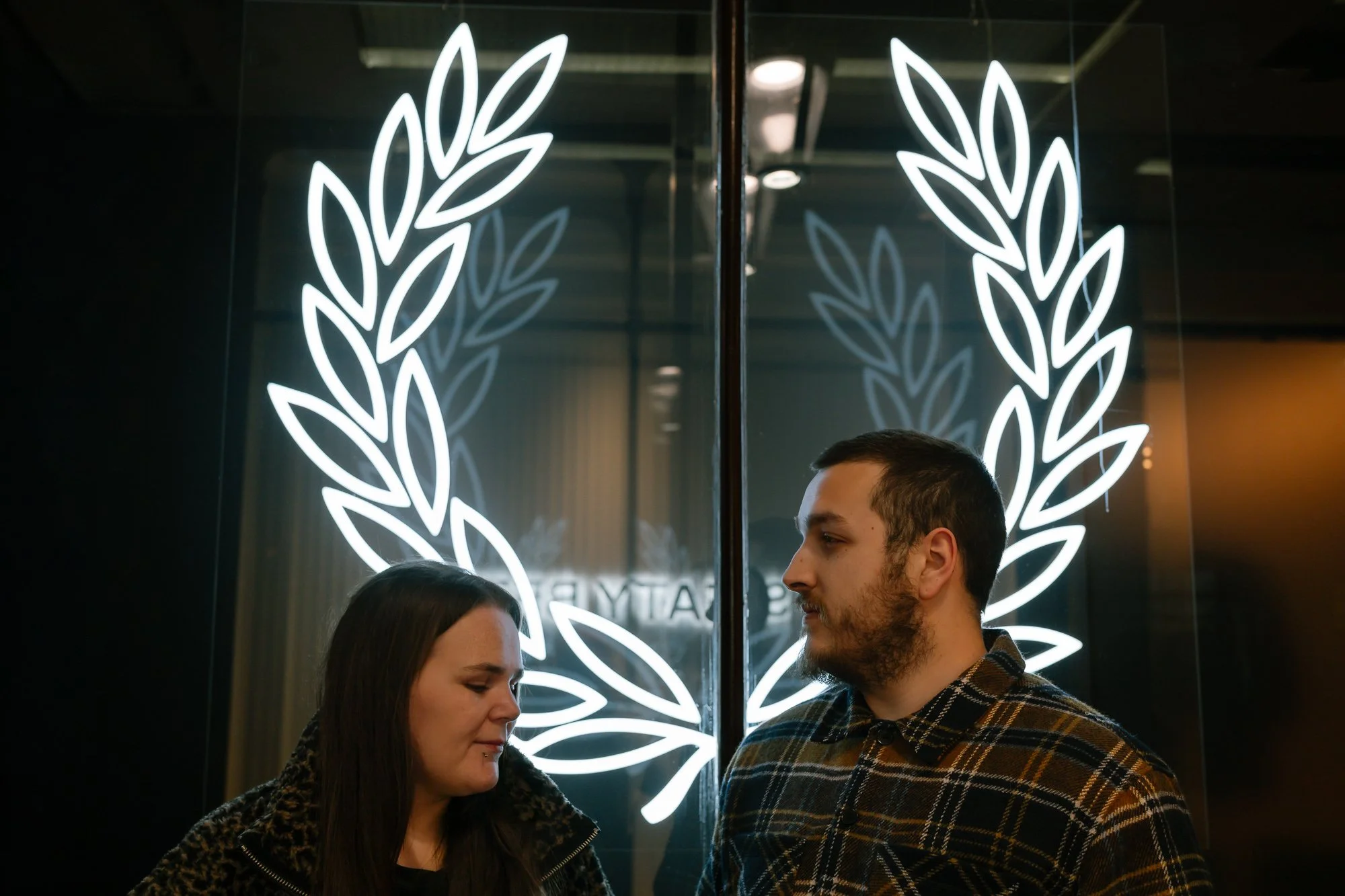 Engaged couple standing beneath glowing neon leaf wreath creating halo effect, romantic urban night portrait in Cardiff