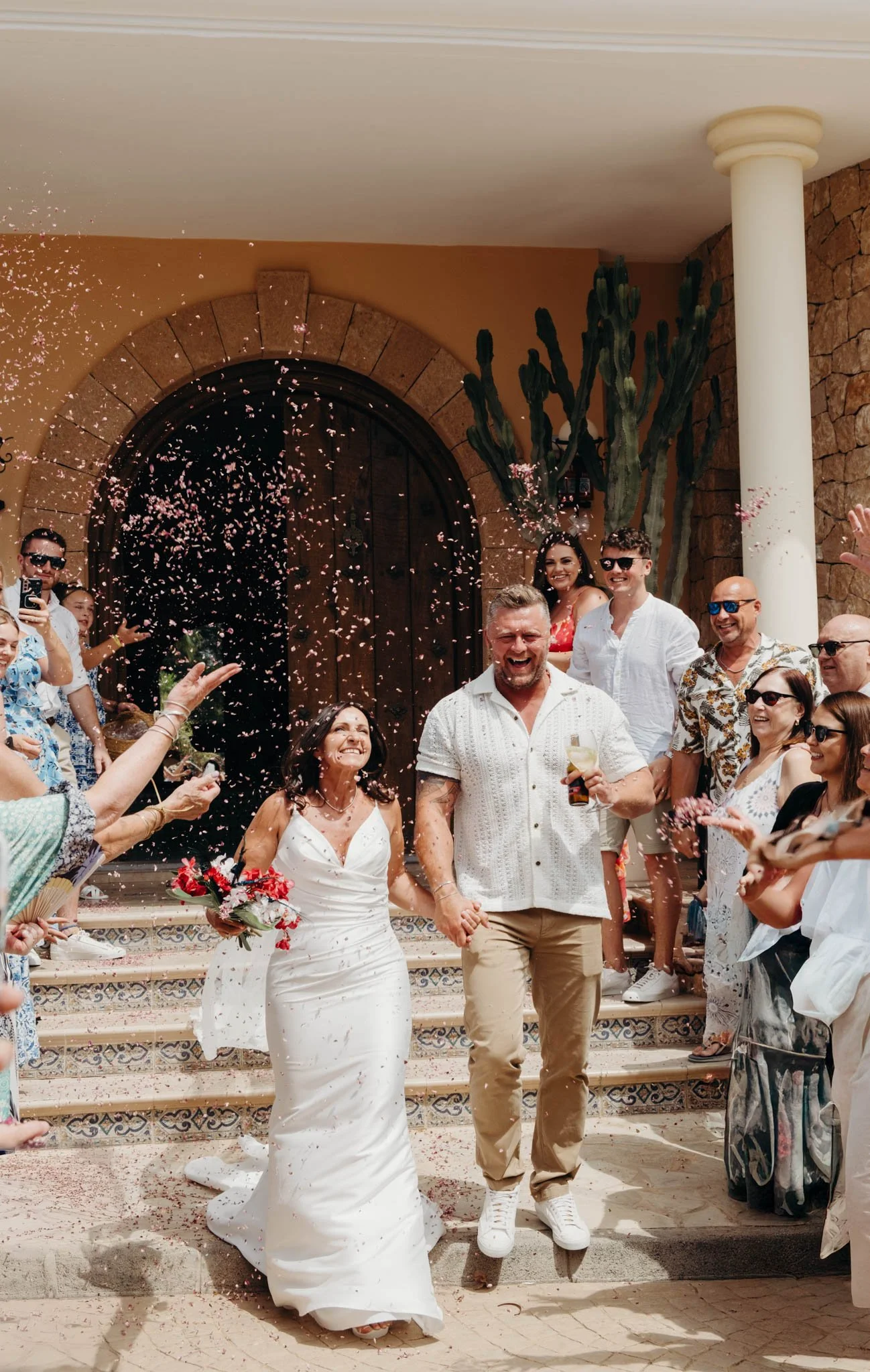 A newly married couple walking down stairs outside a building, surrounded by friends and family throwing flower petals and celebrating.