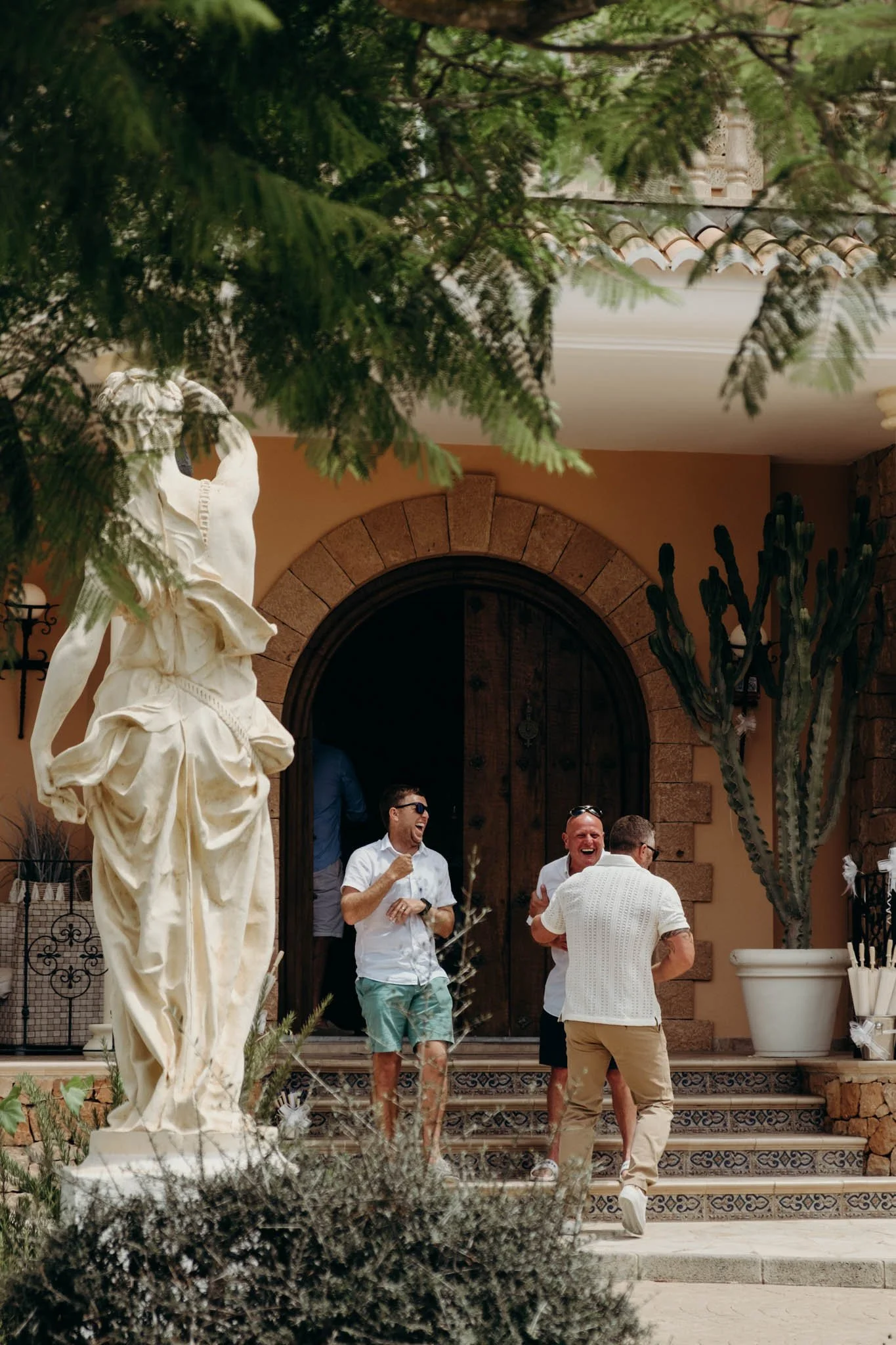 Four men are standing on the steps of a building, smiling and laughing. The building has a large wooden arched door, decorative tiles on the steps, and a large potted cactus. A tree in the foreground partially obscures the scene, and a sculpture of a