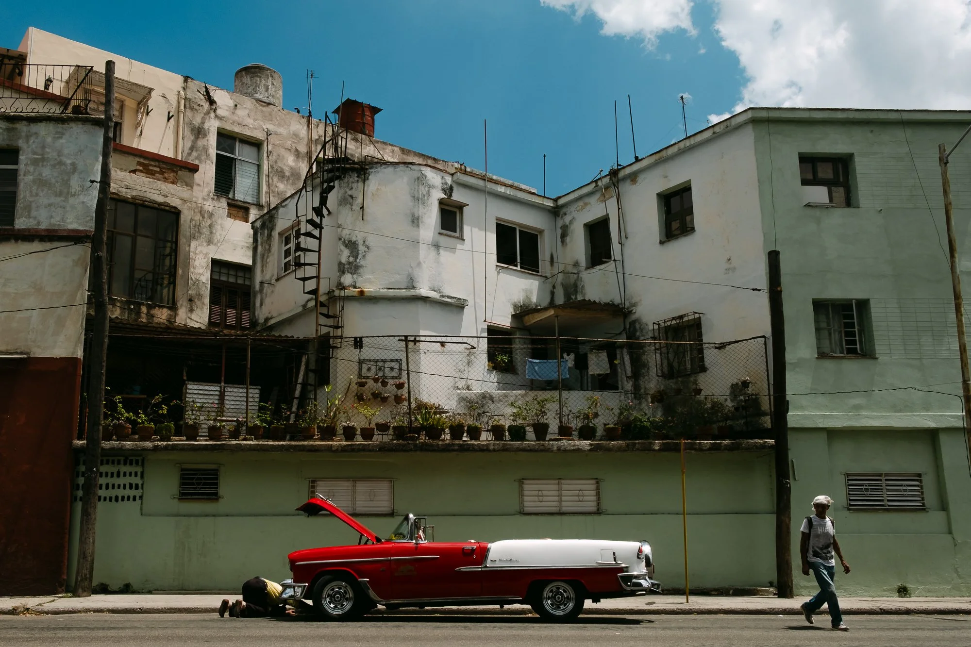A man in Havana on his knees, looking under an old red Cadillac on an empty street, whilst another person crosses the road. 