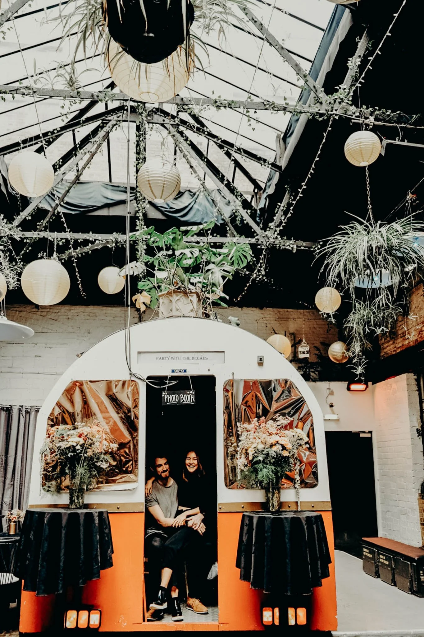 Two people sitting inside a vintage photo booth with floral arrangements on either side, in a decorated indoor space with hanging paper lanterns and plants.