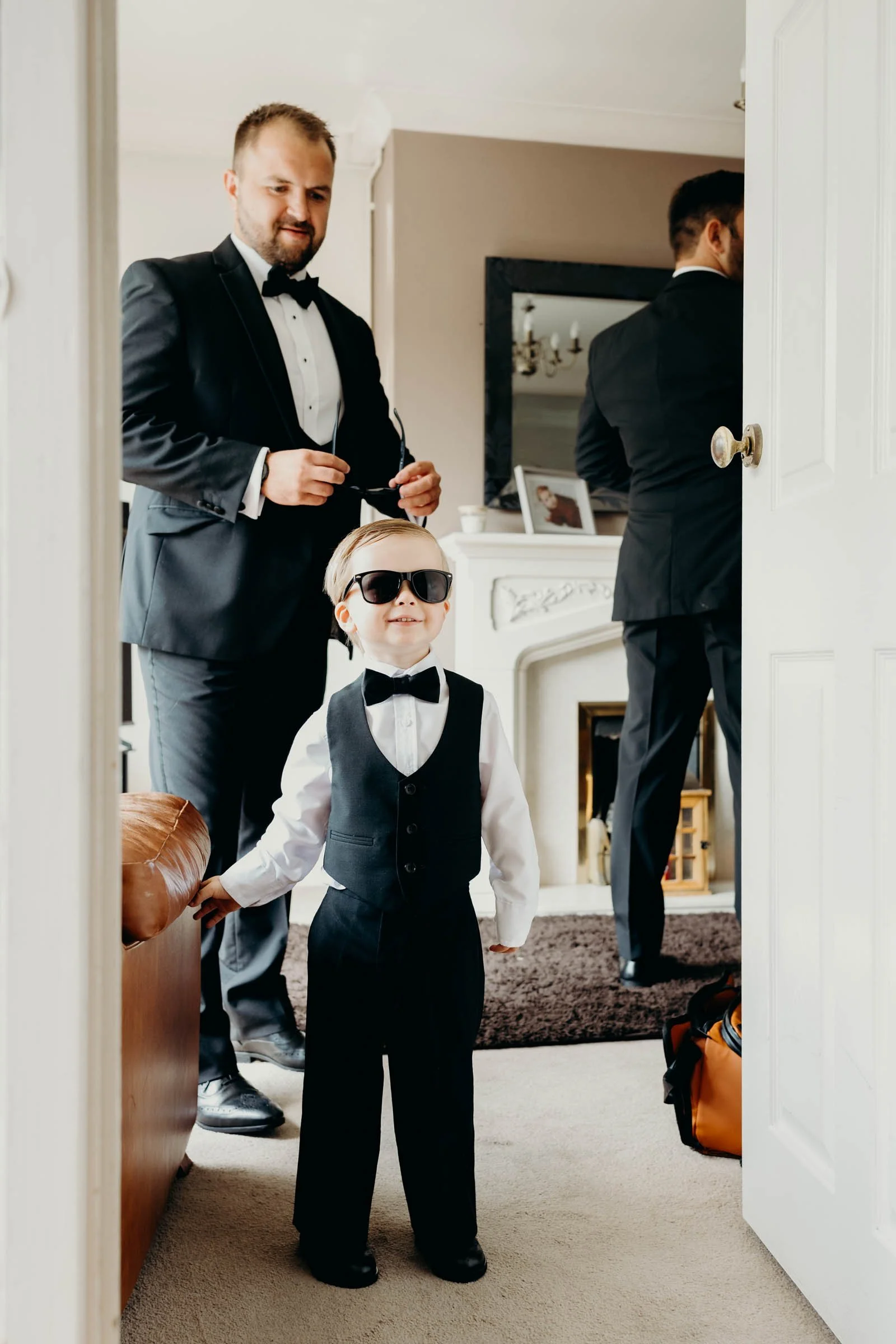 A young boy in a black vest, white shirt, black bow tie, and sunglasses standing in a doorway, smiling, with two men in tuxedos in the background.