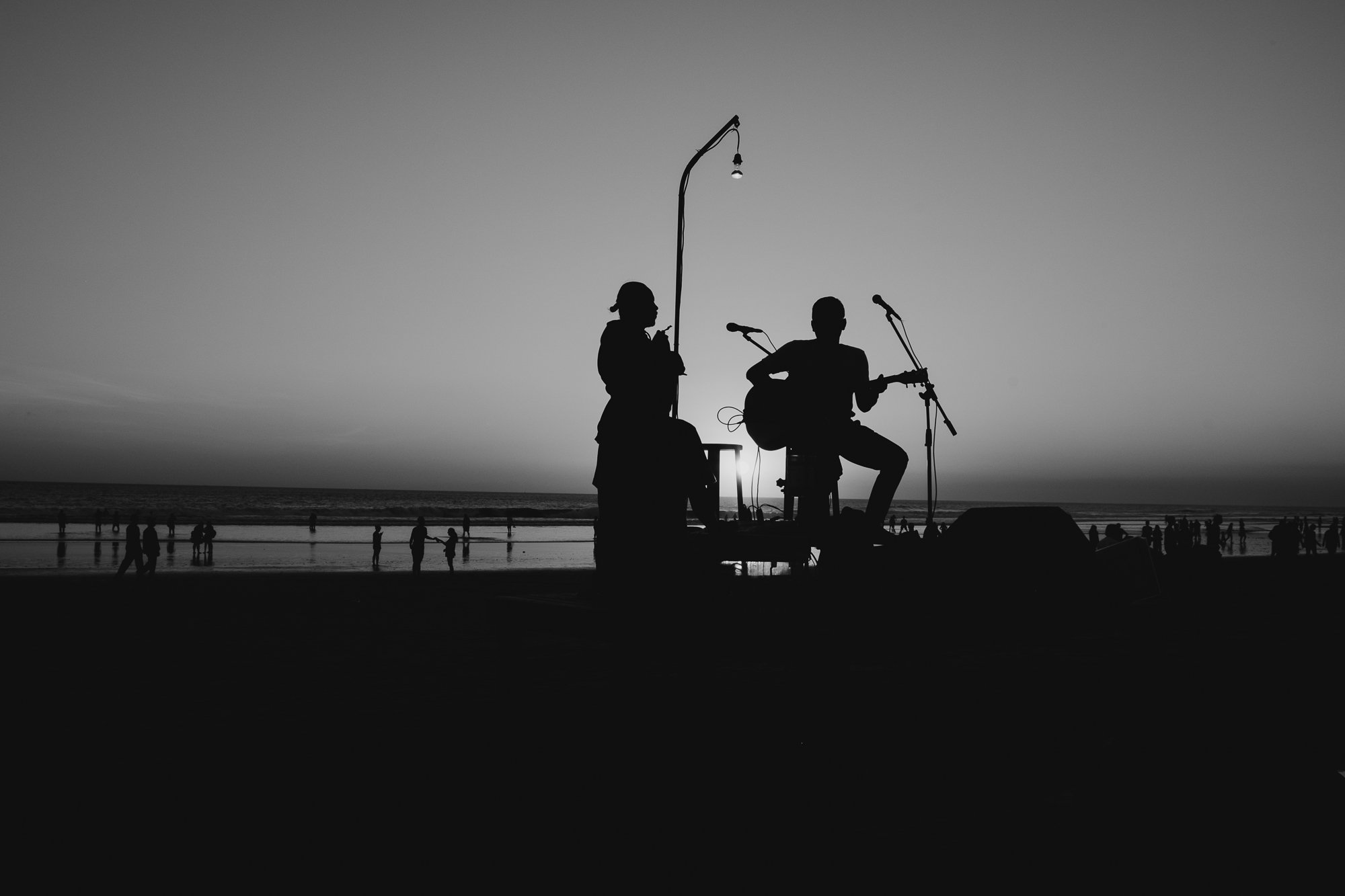 A dramatic black-and-white silhouette in Bali, of two street musicians performing at sunset on a beach: one plays a wind instrument, the other strums a guitar. The low sun creates a bright halo behind them, while scattered silhouettes of people walk 