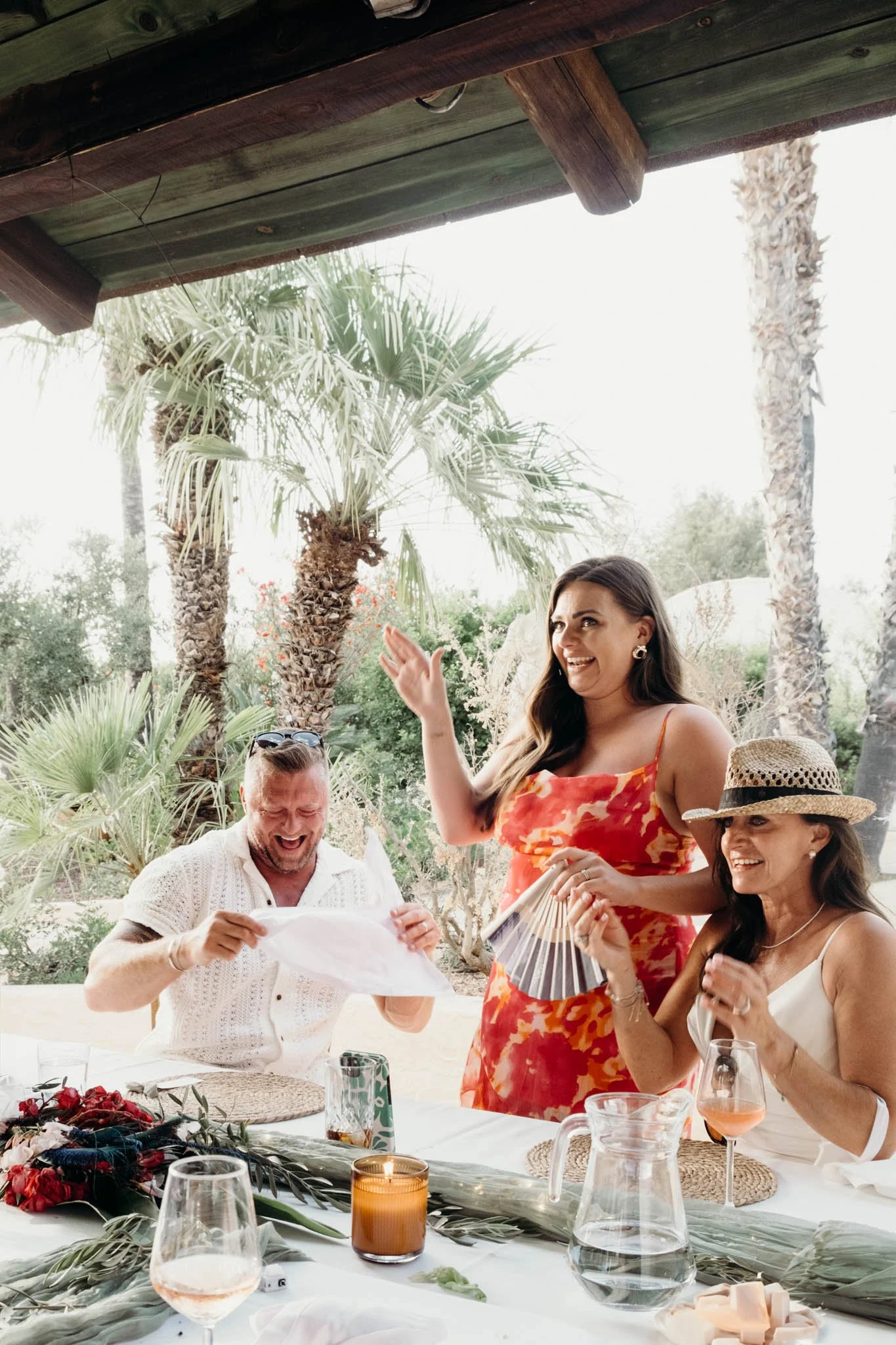 Three women and one man sitting at a table outdoors surrounded by palm trees, celebrating with drinks and laughter.