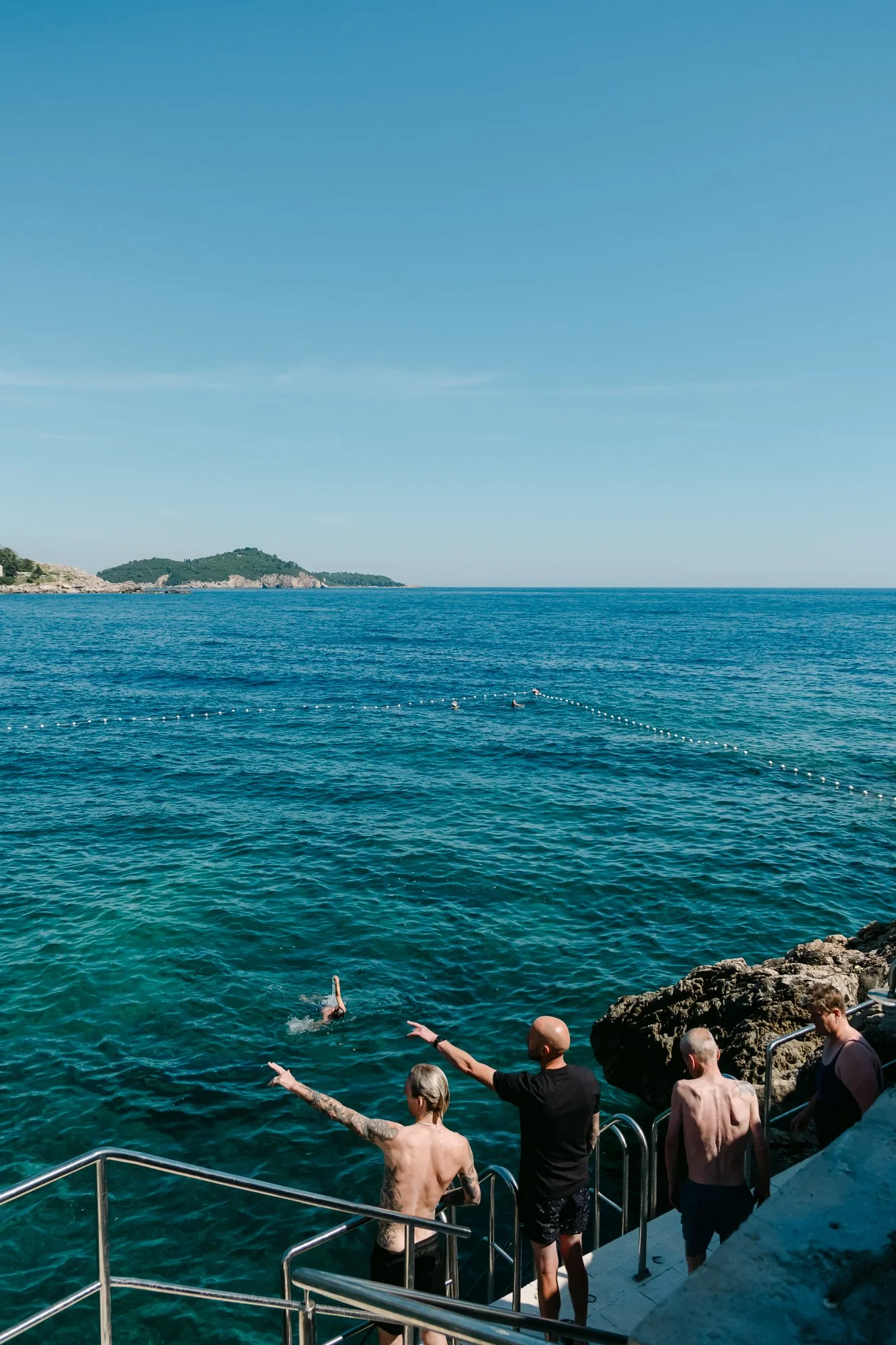 Four shirtless men stand on a concrete platform with metal stairs leading into clear turquoise sea water. Two are pointing toward swimmers in the distance, marked by a buoy line. Rocky cliffs and green islands are visible on the horizon under a brigh