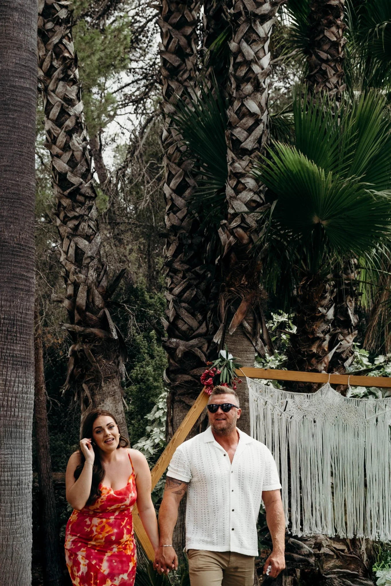 A man and woman hold hands in front of palm trees and a decorated backdrop, outdoors during daylight. The woman wears a colorful dress, and the man wears sunglasses, a white short-sleeved shirt, and beige pants.