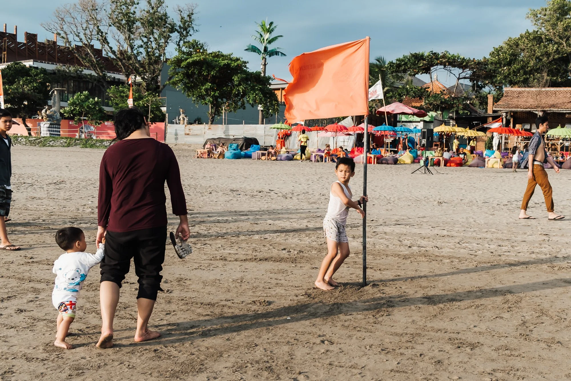 A child on the beach in Bali, holds onto a pole with a red flag in front of a beach bar 