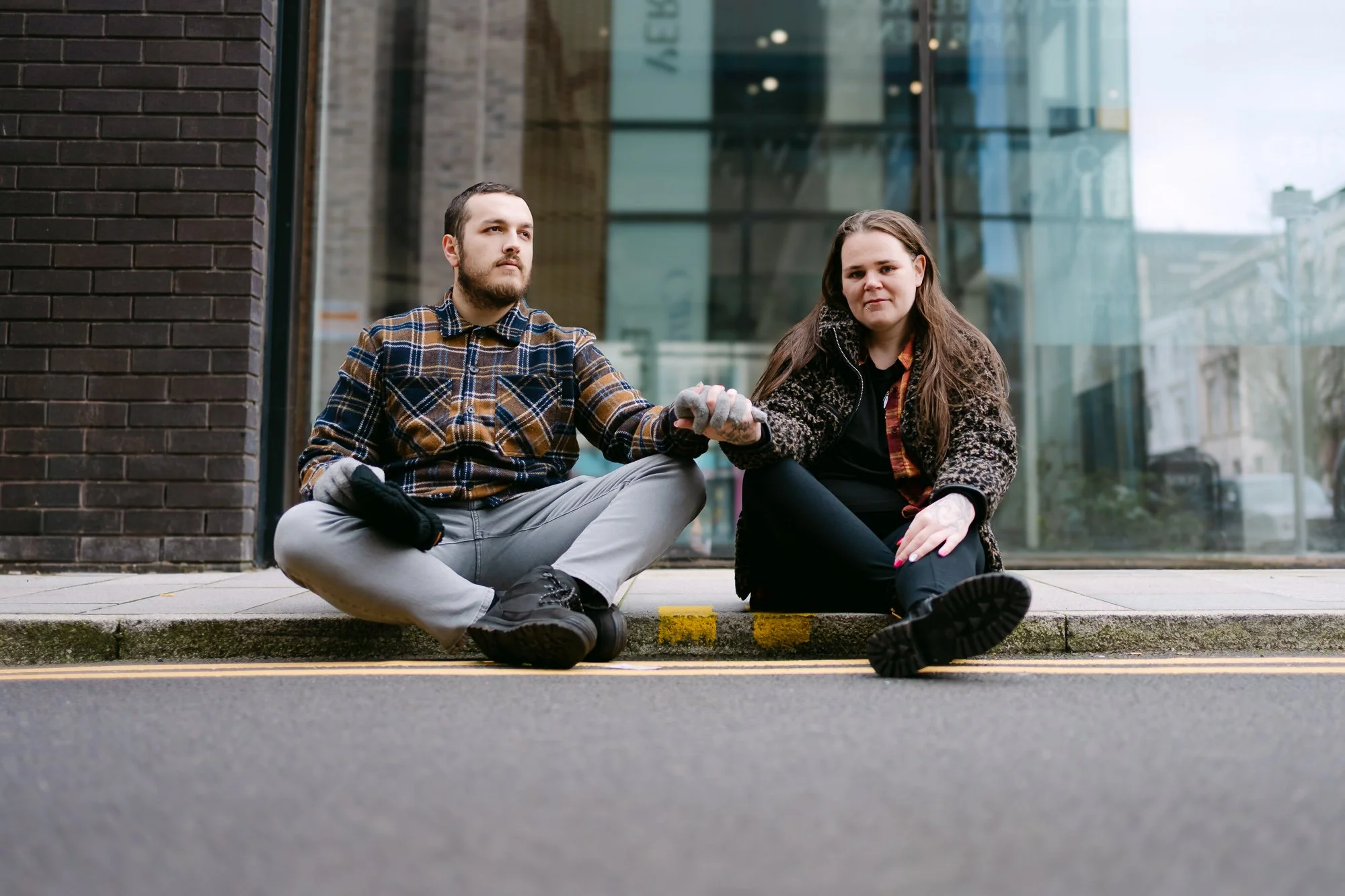 Couple holding hands sitting on a curb being photographed for their engagement in Cardiff 