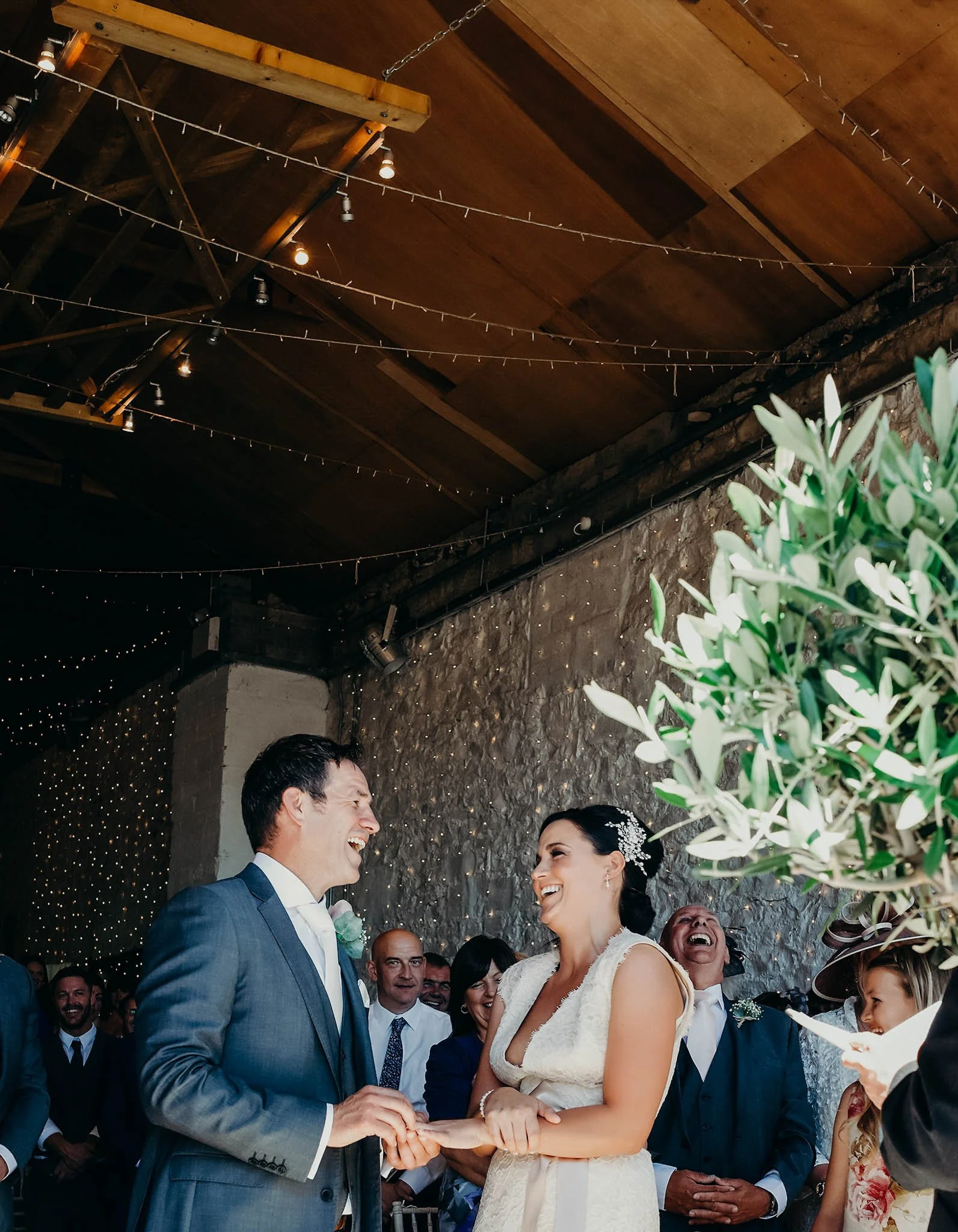 A bride and groom smile at each other during their wedding ceremony, holding hands while exchanging vows, surrounded by friends and family. The setting has a rustic wooden ceiling and string lights.