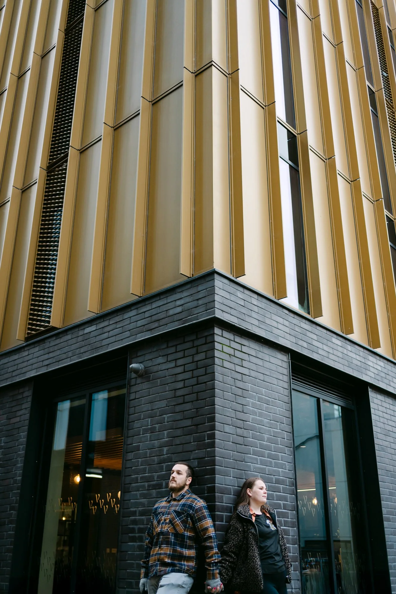Couple secretly holding hands around sleek gold-panelled building corner, contemporary city engagement photo