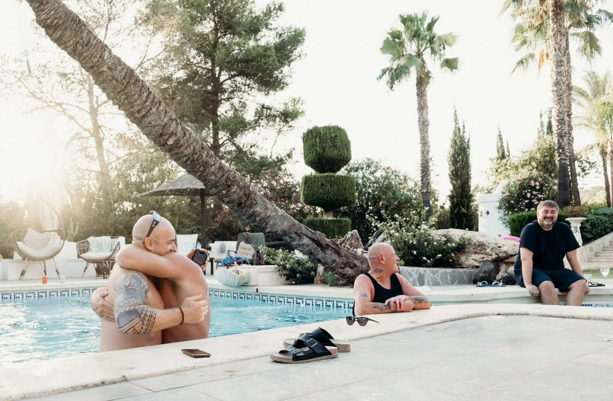 Three men at a backyard swimming pool during sunset, with palm trees and lush greenery in the background. Two men are in the pool hugging, and one man is sitting on the pool's edge, smiling.