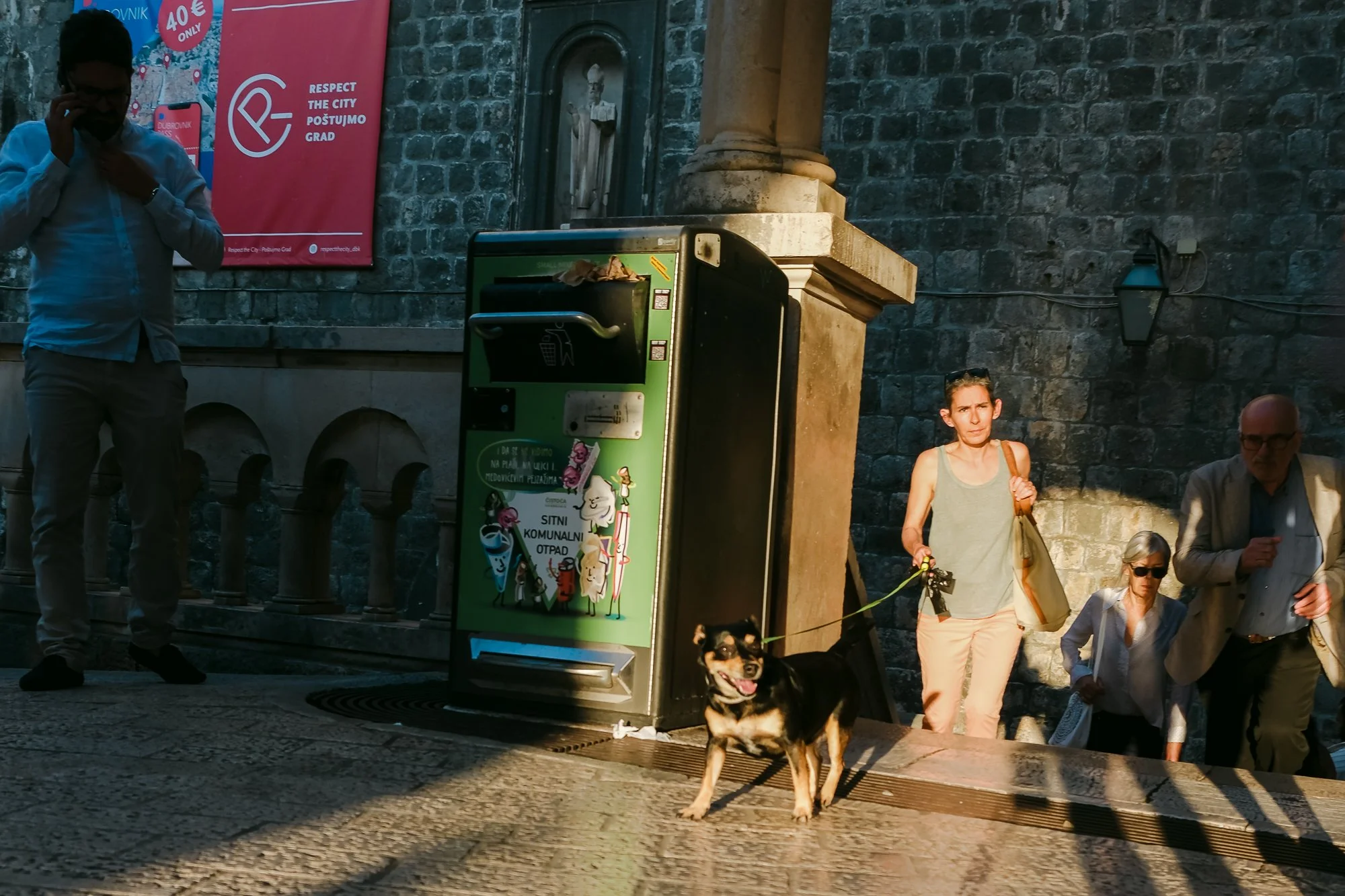 A person walks a happy, tongue-out dog on a leash down stone steps in a historic European old town. Nearby, a green public waste bin is covered in stickers and posters urging proper trash disposal. A red "RESPECT THE CITY" banner hangs on an ancient 