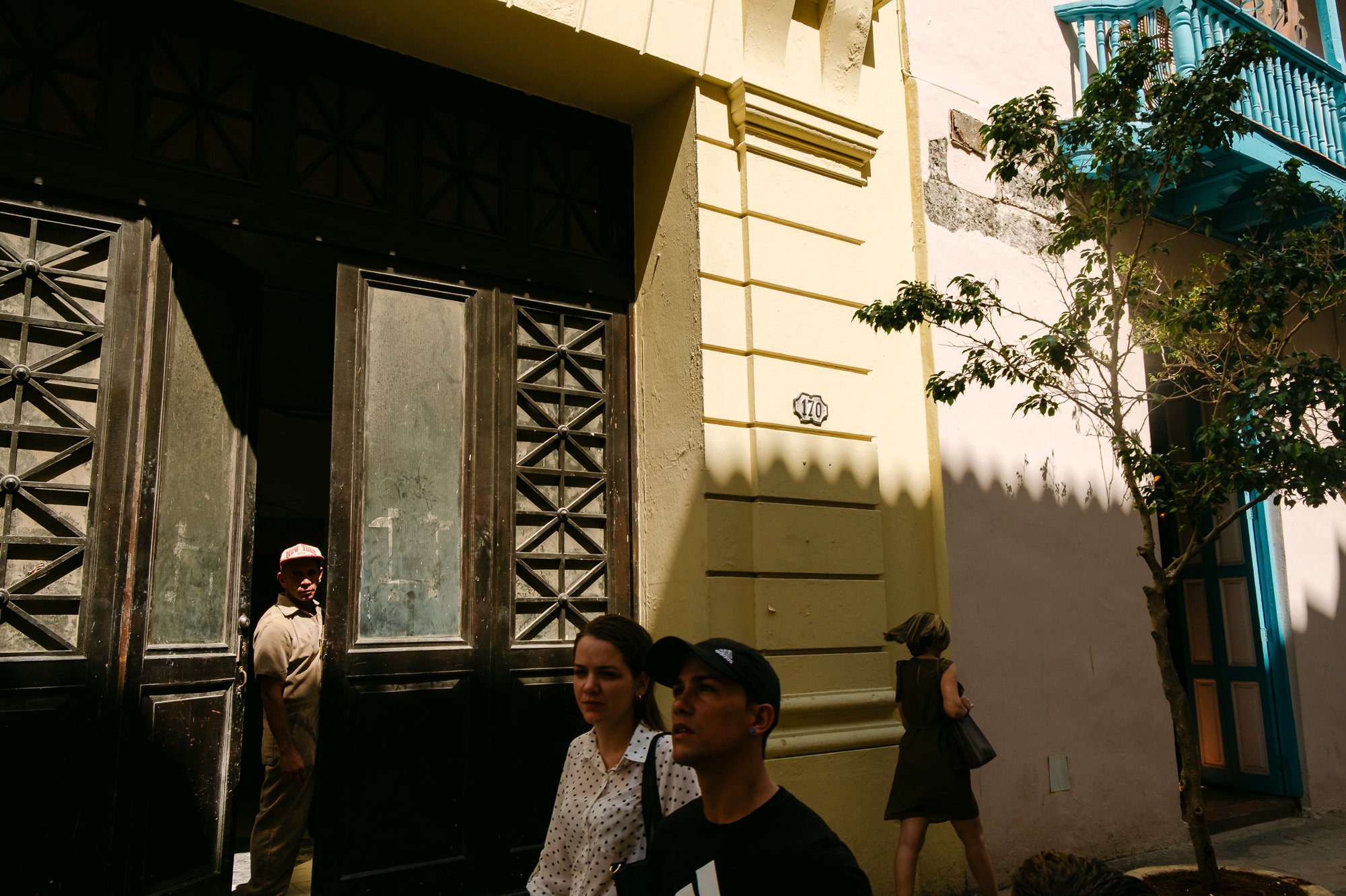 A man in a red-and-white cap stands inside an open ornate metal gate of a historic building, looking out toward the street. In the foreground, a young couple (woman in a polka-dot blouse, man in a cap) walks past a pale yellow colonial-style facade m