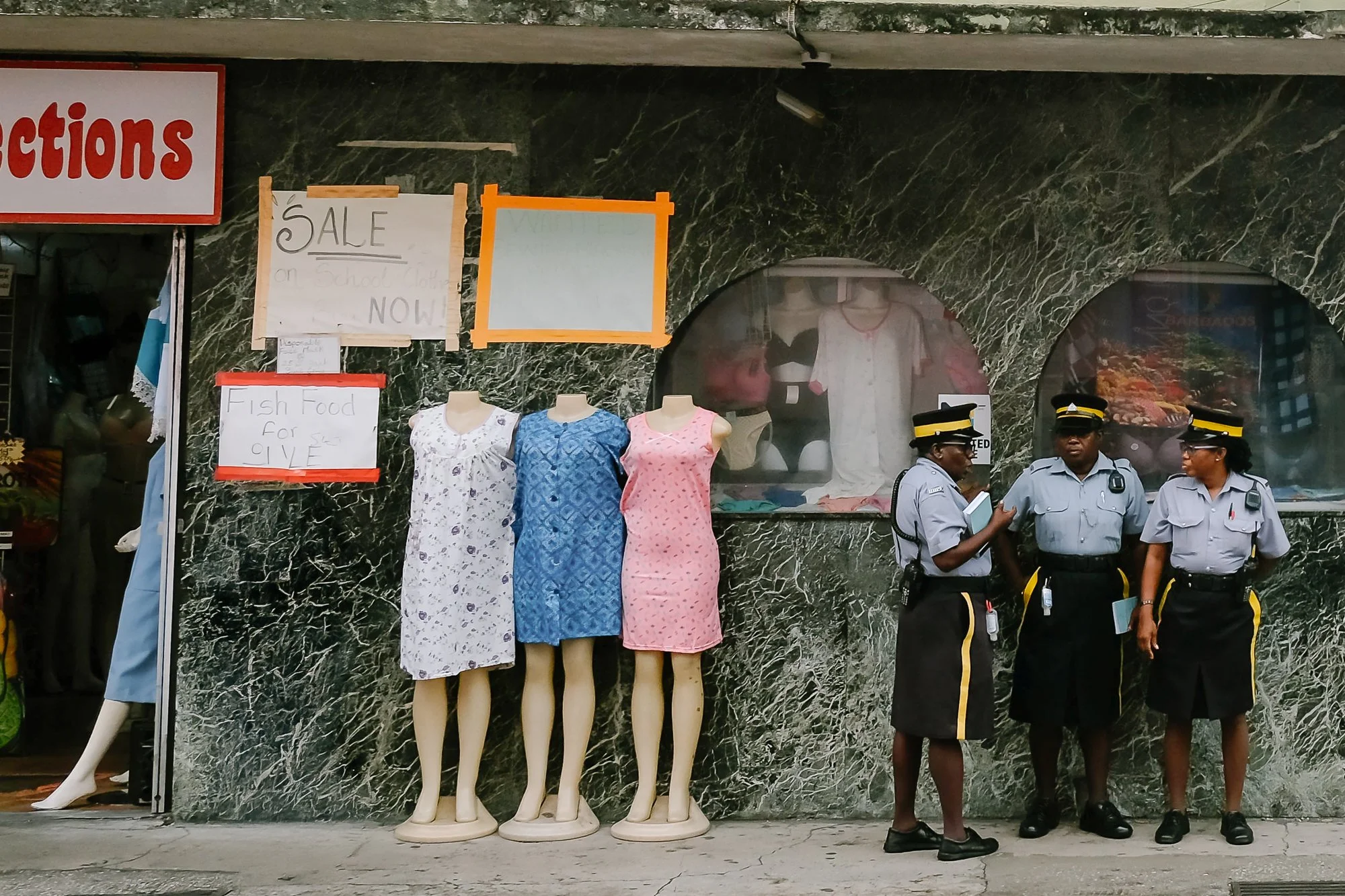 Three headless mannequins in Bridgetown, Barbados, display colorful nightgowns (white floral, blue patterned, and pink polka-dot) outside a shop with a green marble facade. Nearby, three police officers in light blue uniforms stand chatting. Handwrit