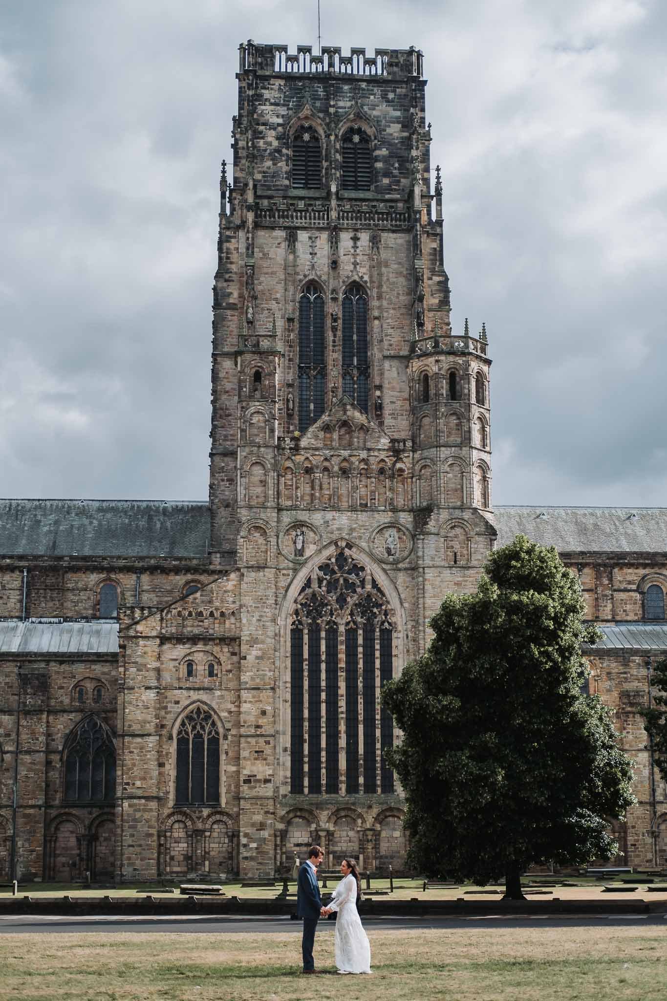 A bride and groom holding hands in front of Durham Cathedral. The sky is cloudy, and there are trees and benches in the foreground in Durham.