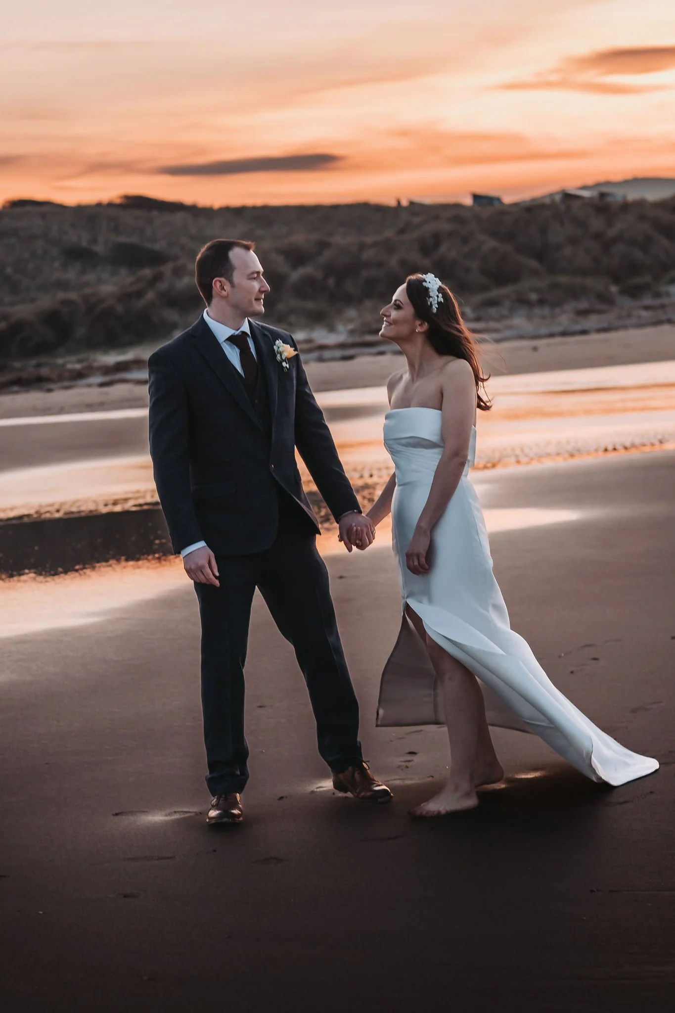 A bride and groom holding hands on a beach at sunset, smiling at each other, with surf and hills in the background.