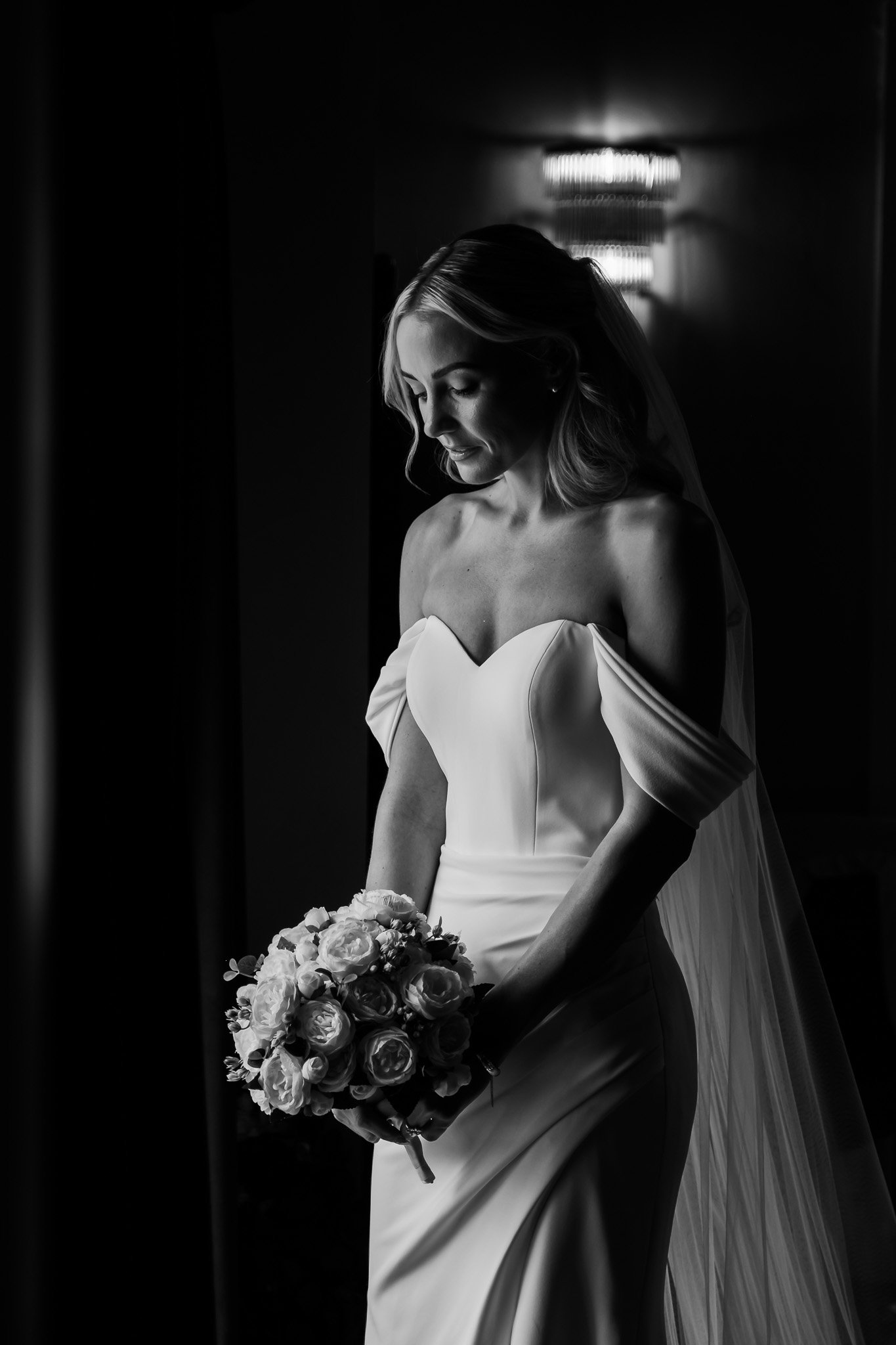 Black and white photo of a woman in a wedding dress holding a bouquet of flowers, standing indoors near a wall-mounted light fixture.