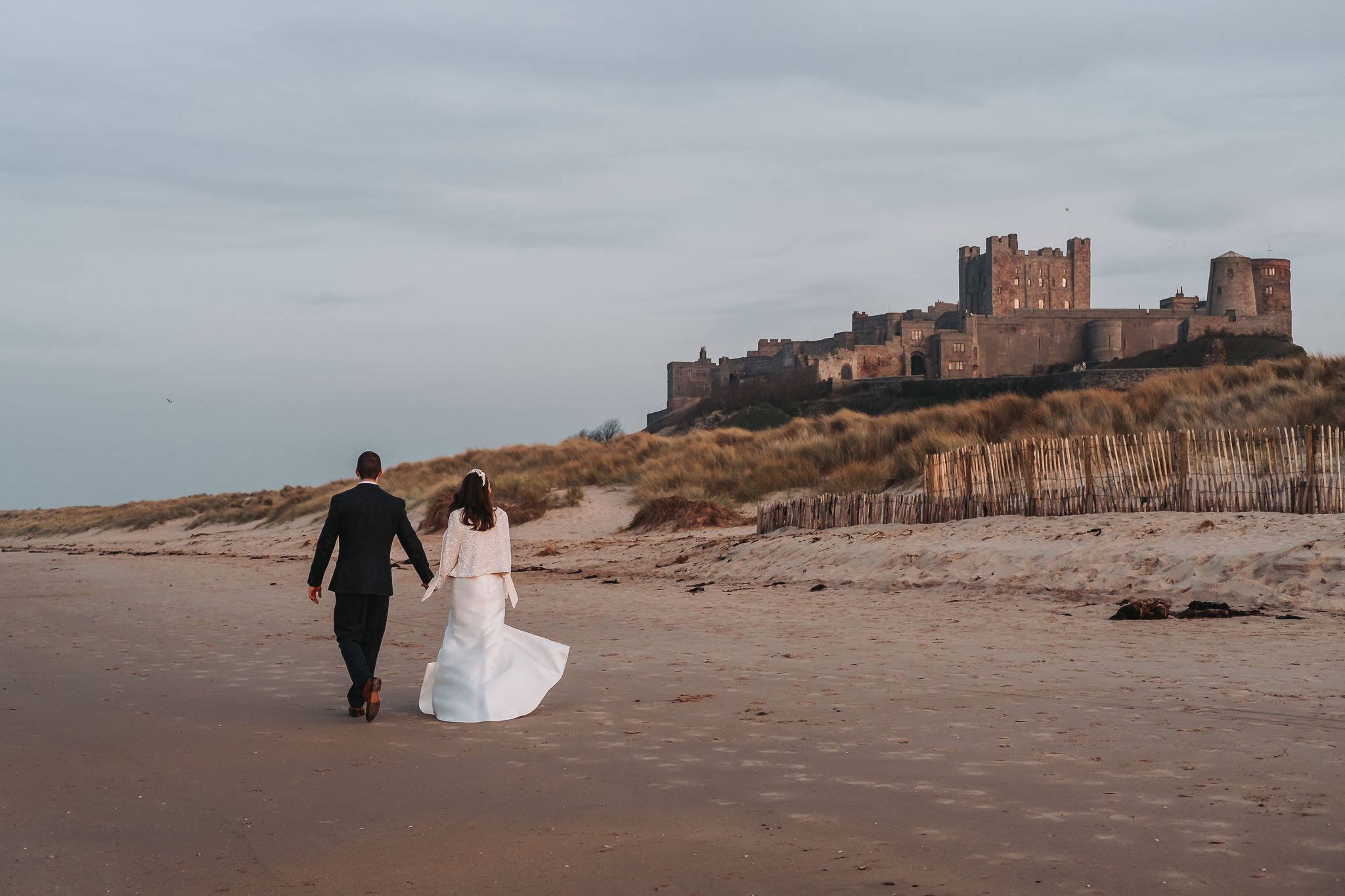 A bride and groom walking hand in hand on a deserted beach near Bamburgh Castle in the distance under a cloudy sky.