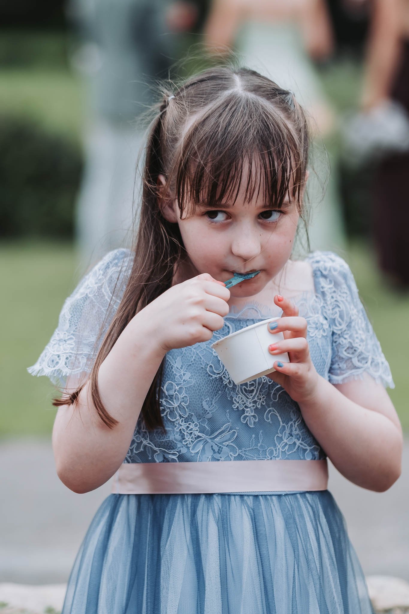 A young girl with wet hair wearing a light blue dress with lace details, eating ice cream from a white cup outdoors.
