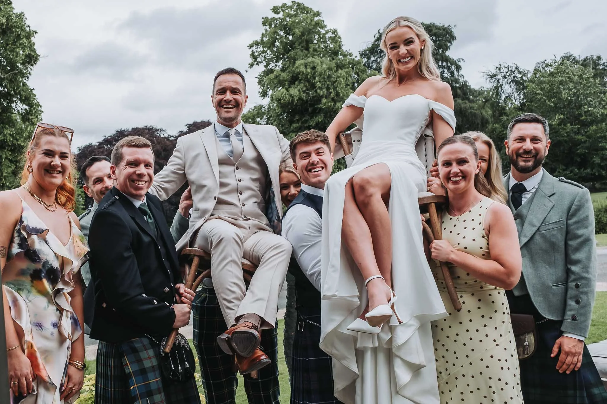 Group of people outdoors at a wedding celebration, with a woman in a white dress being lifted on a chair, surrounded by smiling friends and family at Whitworth Hall.