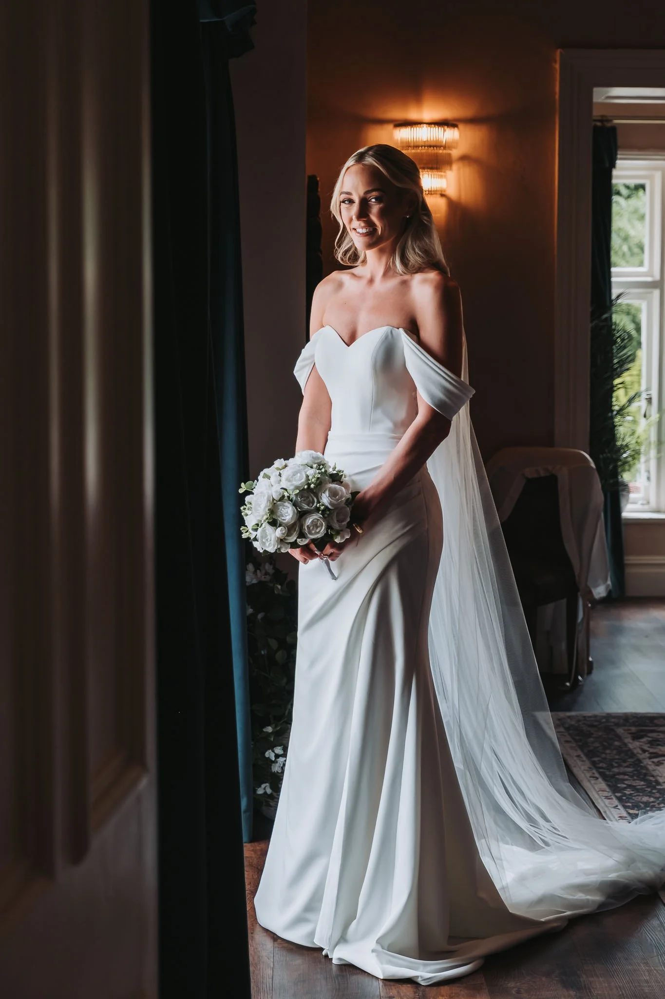 A smiling bride in a white wedding dress holding a bouquet of white roses, standing indoors near a window with natural light and wall-mounted light fixtures, with dark curtains and facing the camera at Whitworth Hall.