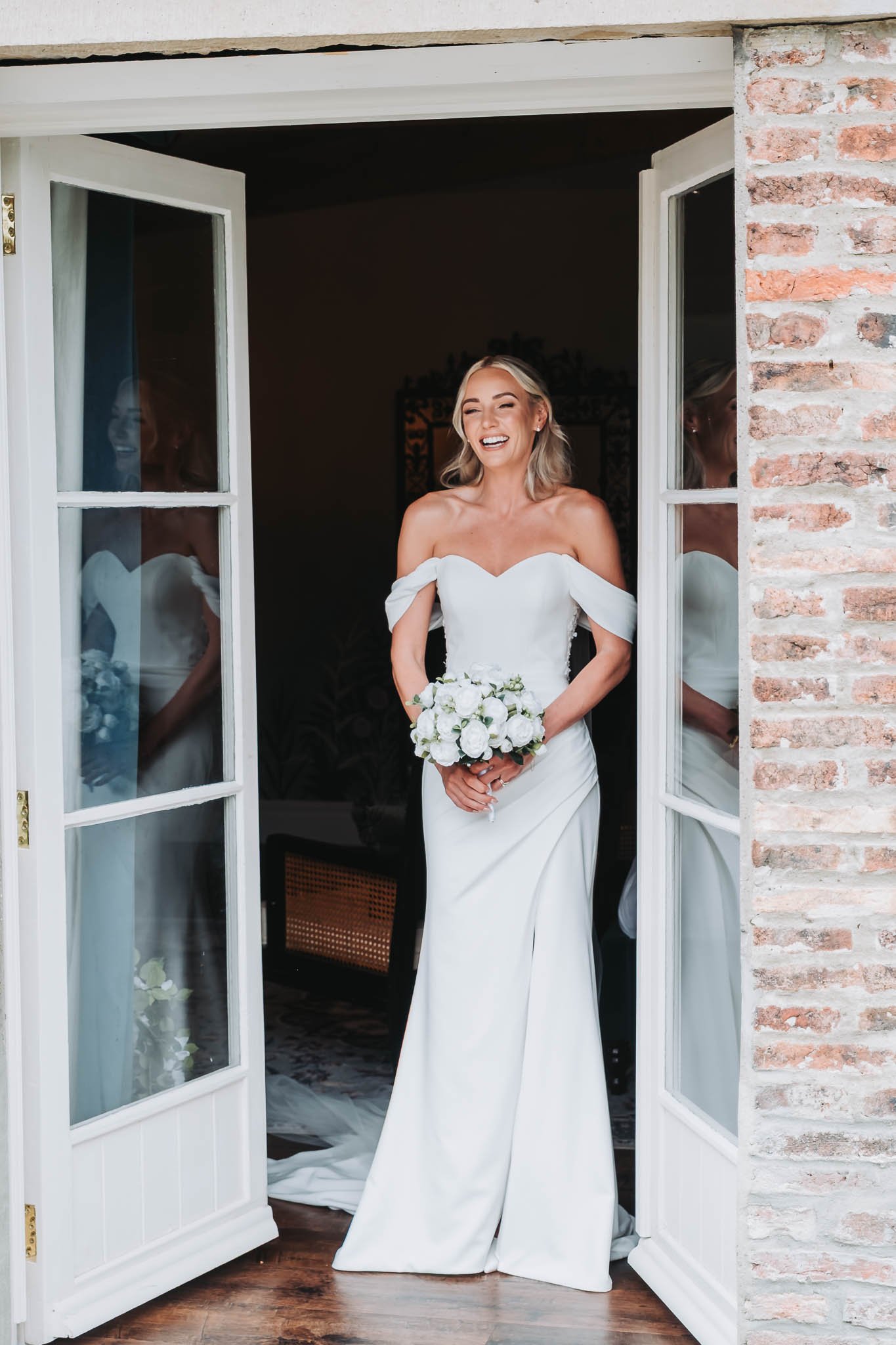 A bride with blonde hair in a white wedding dress holding a bouquet of white flowers, standing in a doorway with a brick wall on the side, smiling.