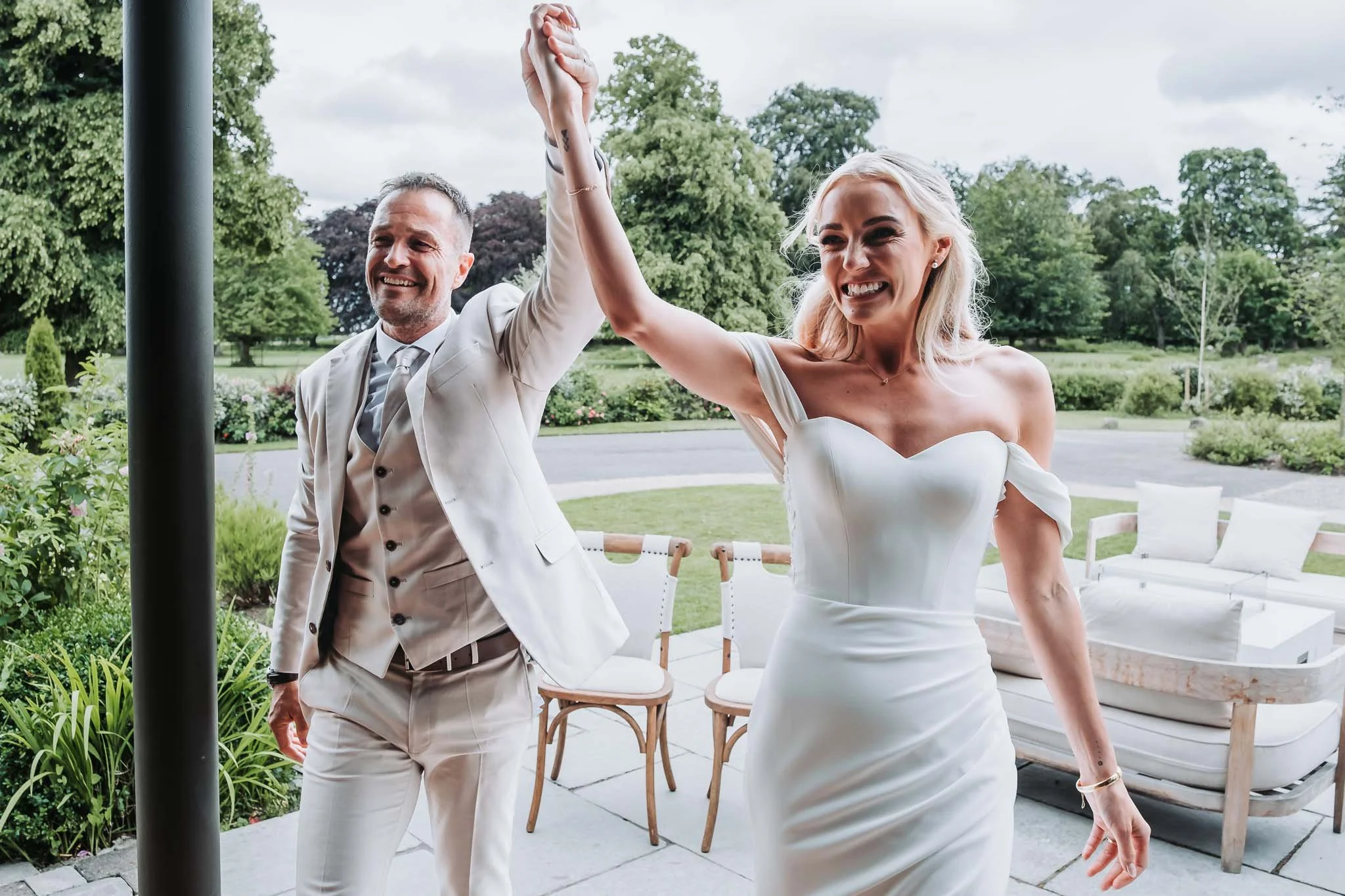A man and woman in wedding attire holding hands and smiling outdoors with trees and a cloudy sky in the background at Whitworth Hall.