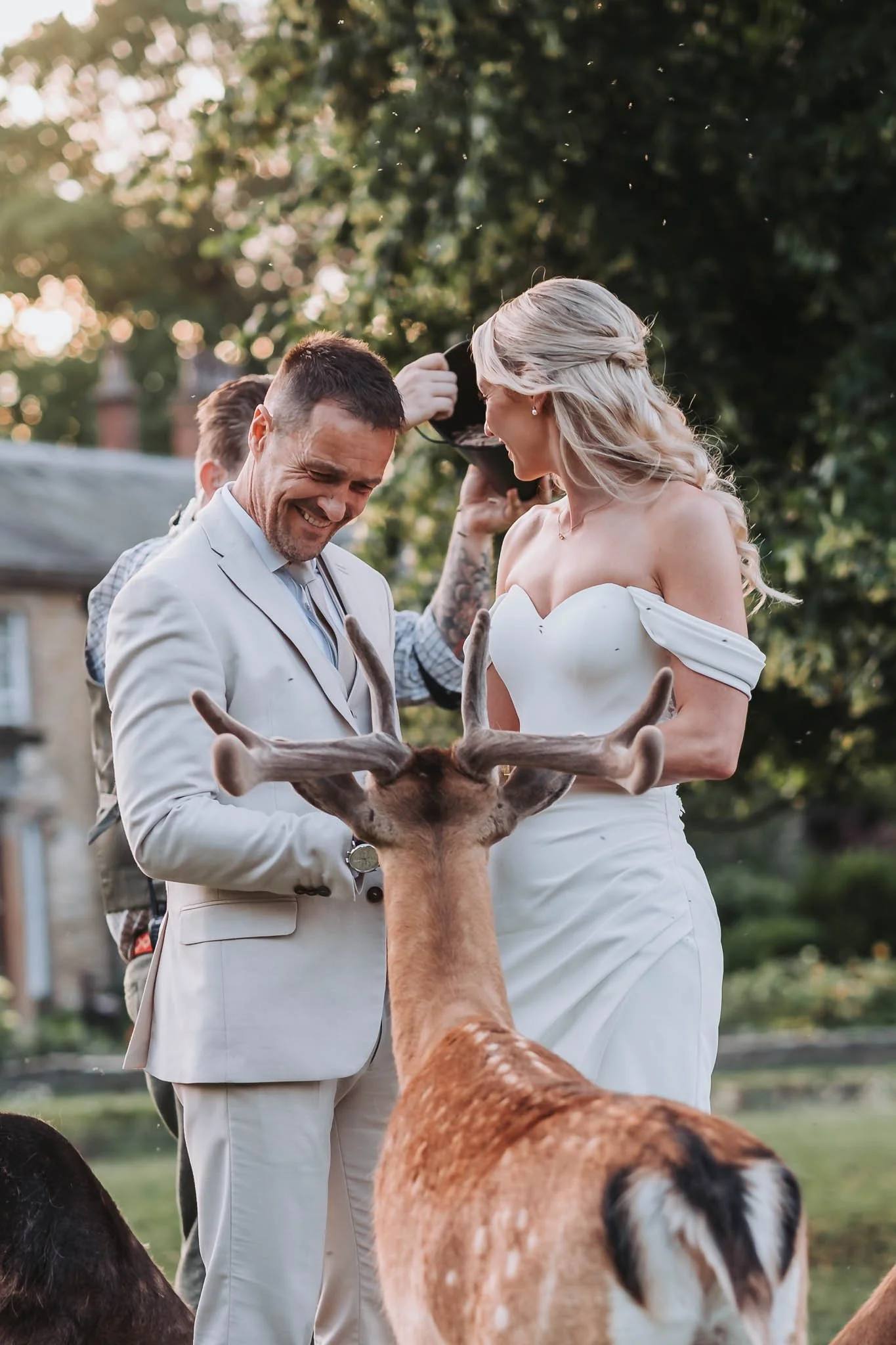 A groom and bride smiling and laughing outdoors, holding a large deer antlers, surrounded by other people and reindeer at Whitworth Hall during daytime.