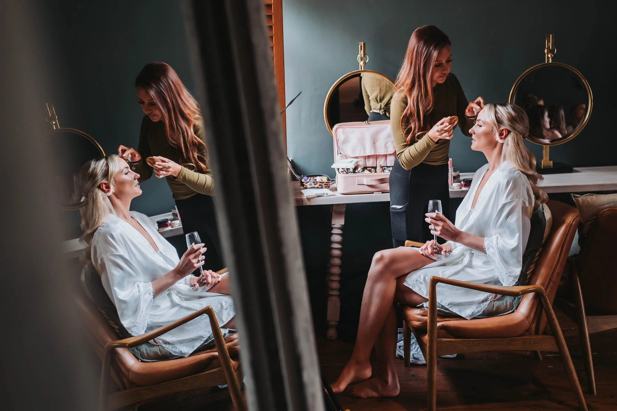 A woman with long, wavy blonde hair sits on a chair, wearing a white robe and holding a glass of wine, while another woman with long, brown hair styles her hair in a salon or dressing room with mirrors and makeup supplies visible in the background.