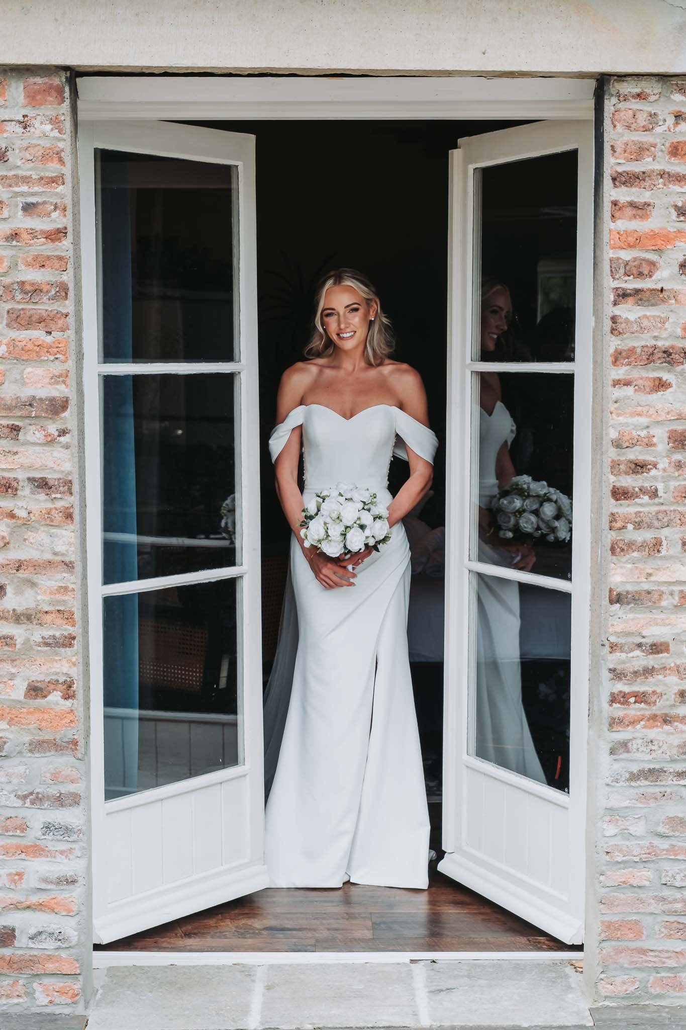 A bride in a white wedding dress holding a bouquet of white roses, standing in an open window with a brick wall at Whitworth Hall.