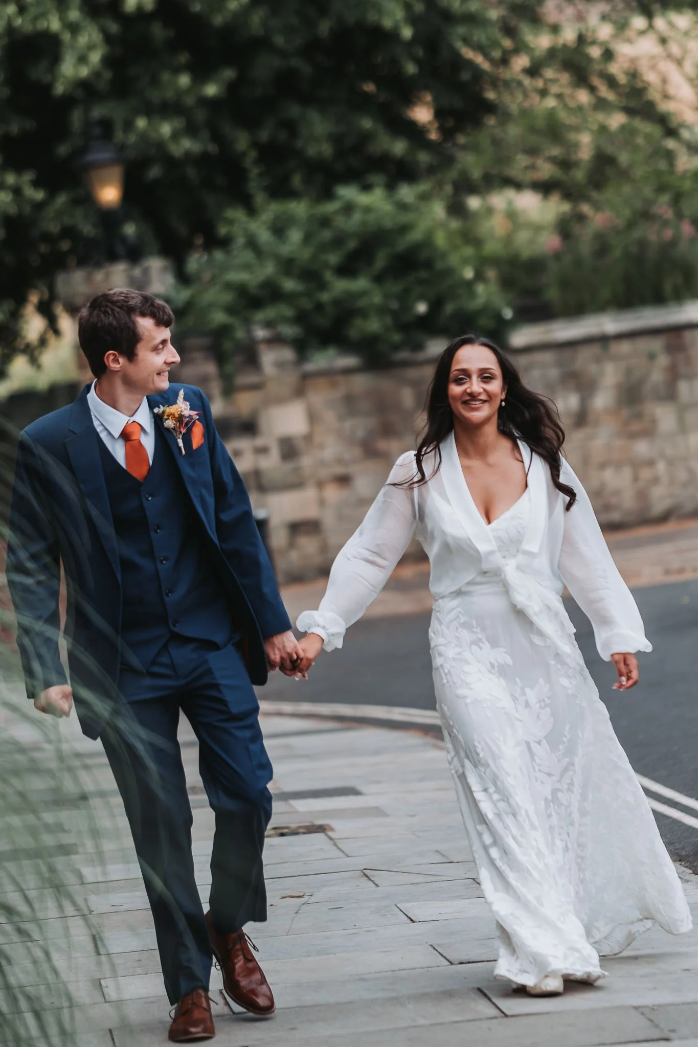 A couple dressed in wedding attire walking hand in hand outdoors. The man is wearing a dark blue suit, white shirt, orange tie, and brown shoes. The woman is wearing a white wedding dress with floral embroidery and long sleeves. They are smiling and 