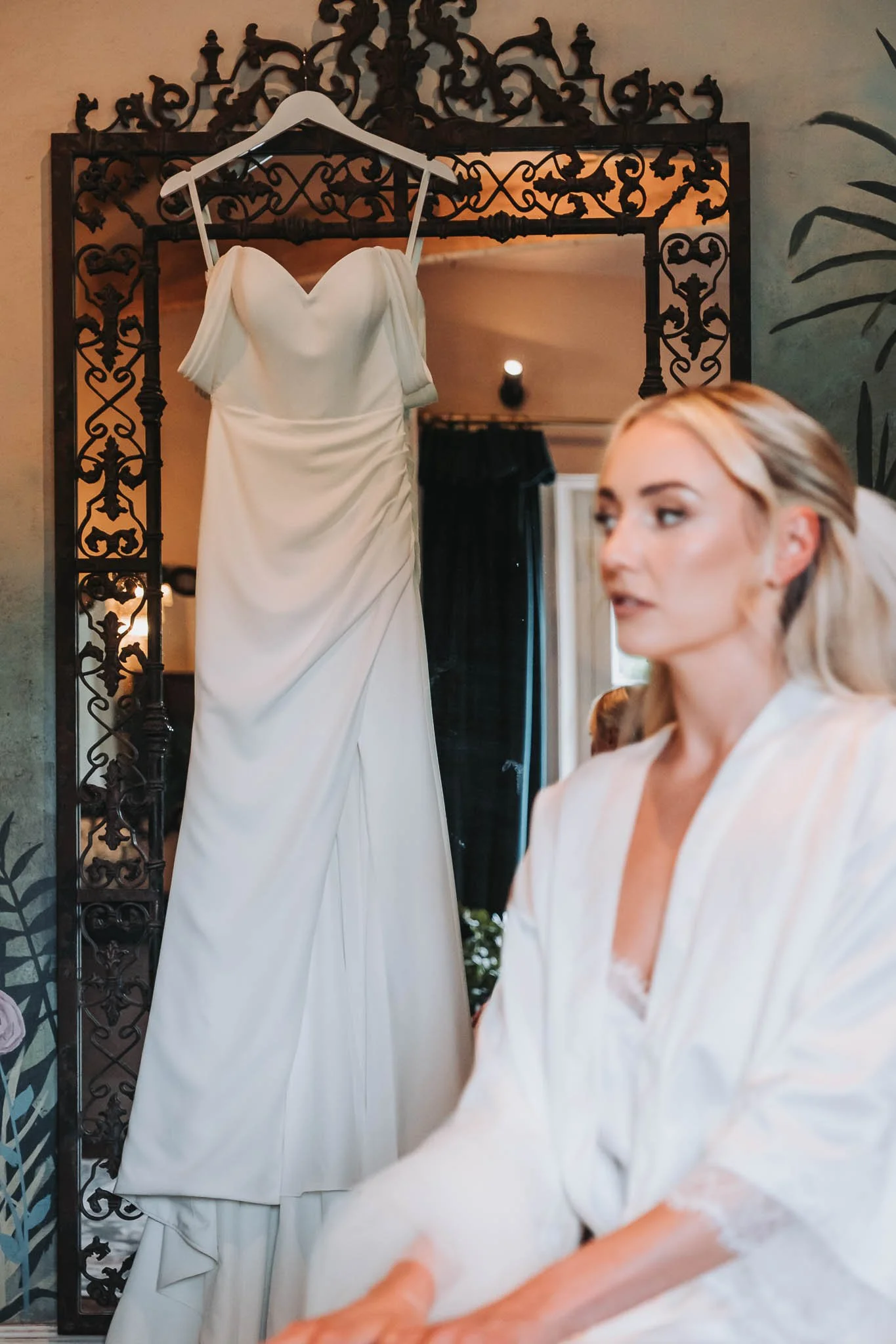 A wedding dress is hanging on a decorative black iron frame, with a woman in a satin robe sitting nearby, preparing for her wedding or fitting.
