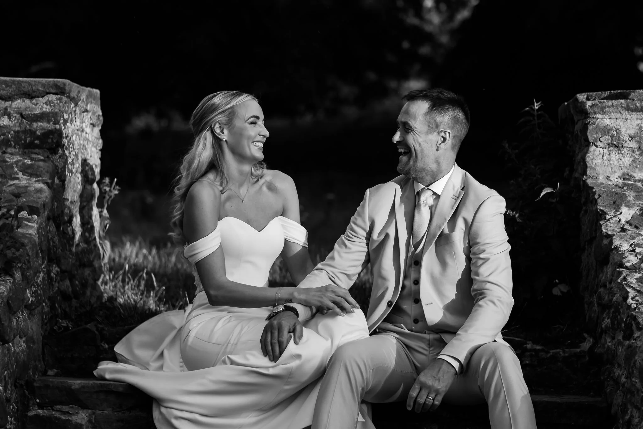 A black-and-white photo of a smiling woman in a wedding dress holding hands with a man in a suit, sitting outdoors between two stone pillars, with trees in the background at Whitworth Hall.