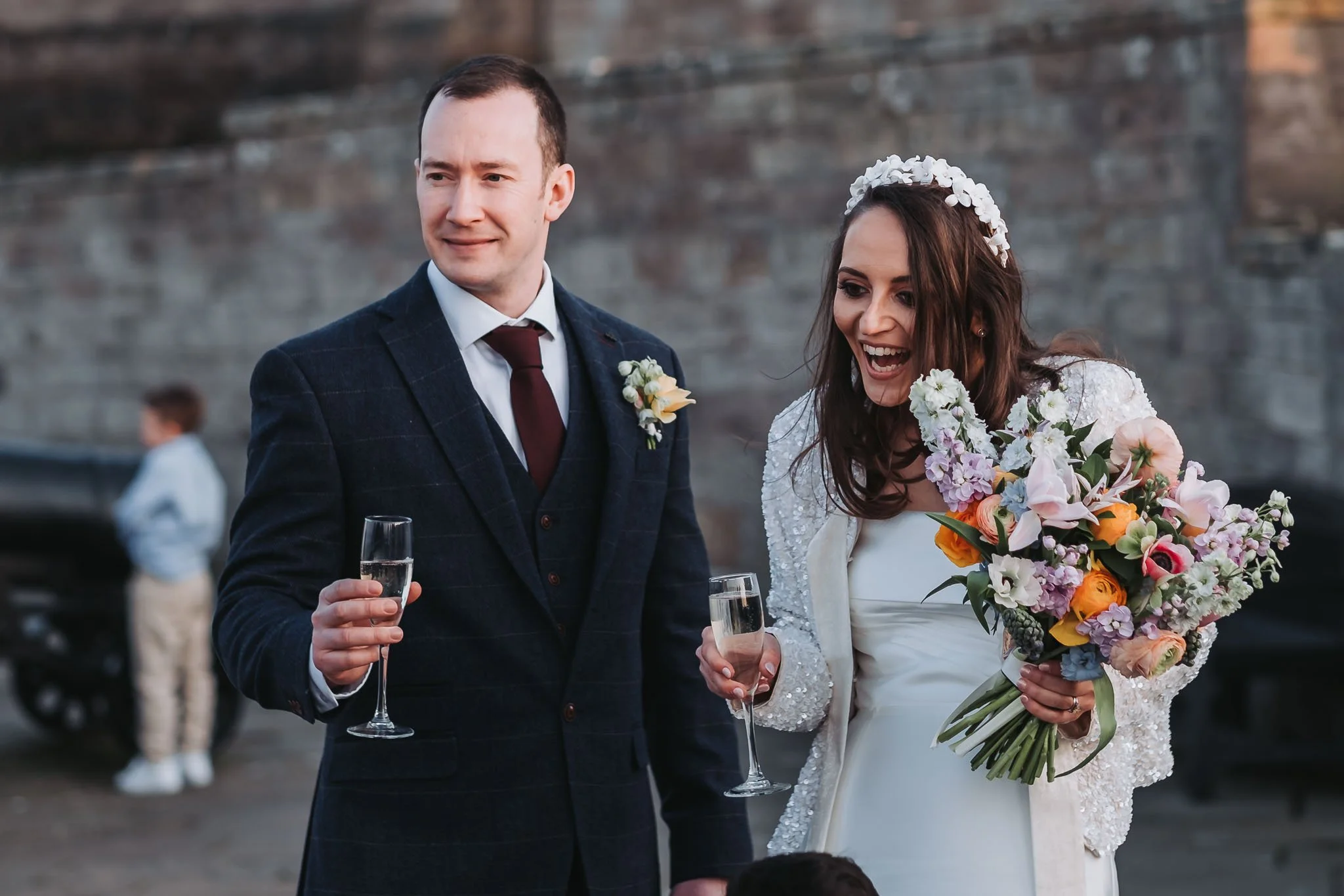 Bride and groom celebrating with glasses of champagne at a wedding reception outdoors, with Bamburgh Castle in the background.
