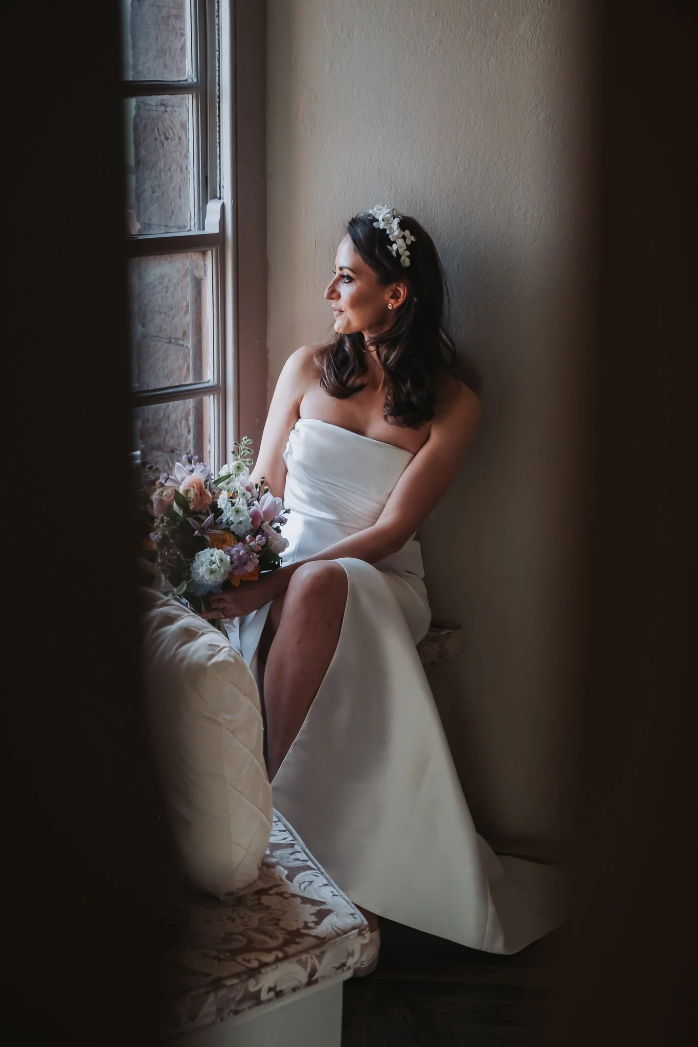 A bride sitting by a window in a white wedding dress holding a bouquet of flowers, looking out contemplatively.