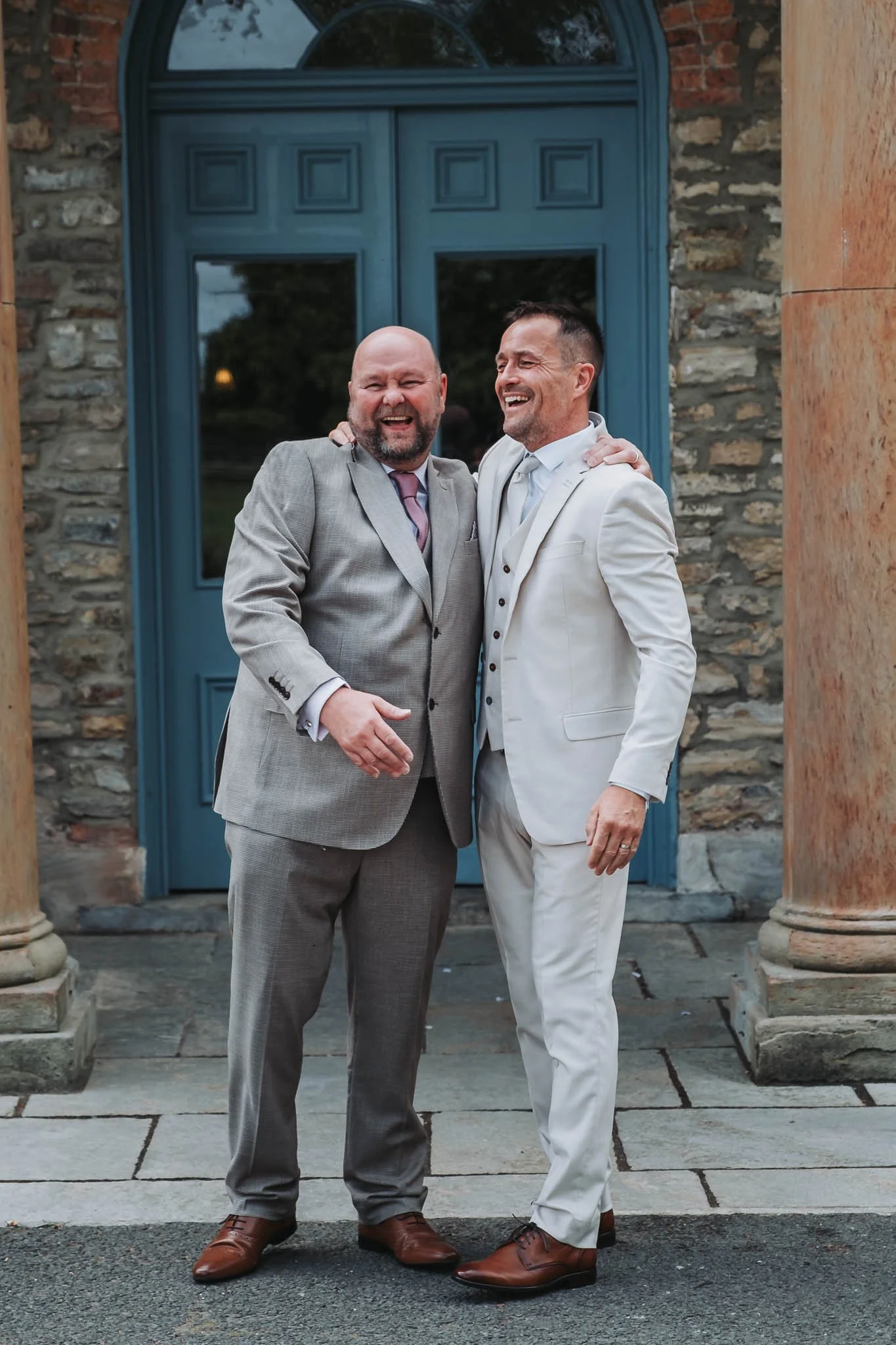 Two men dressed in suits standing close together and smiling, outside a building with stone walls, columns, and a blue door in the background.